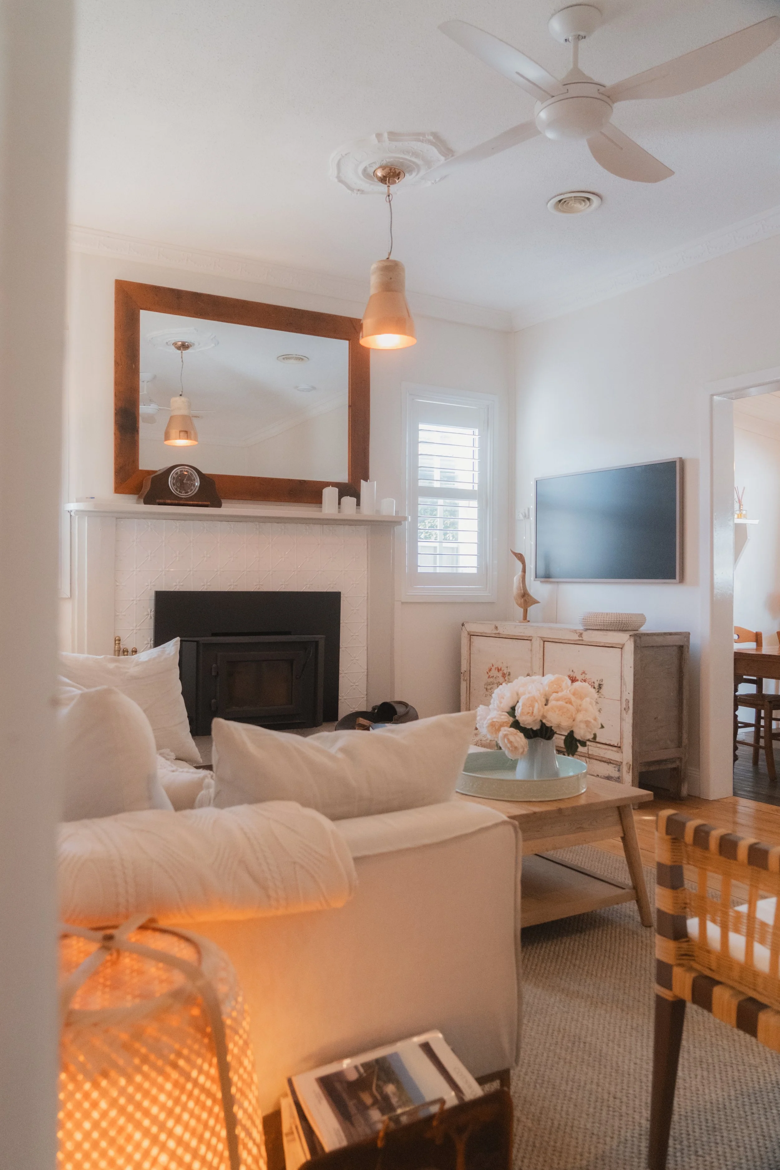 Living room with white sofa, coffee table with flowers, wall-mounted TV, mirror, ceiling fan, pendant light, window with blinds, and various decorative items.