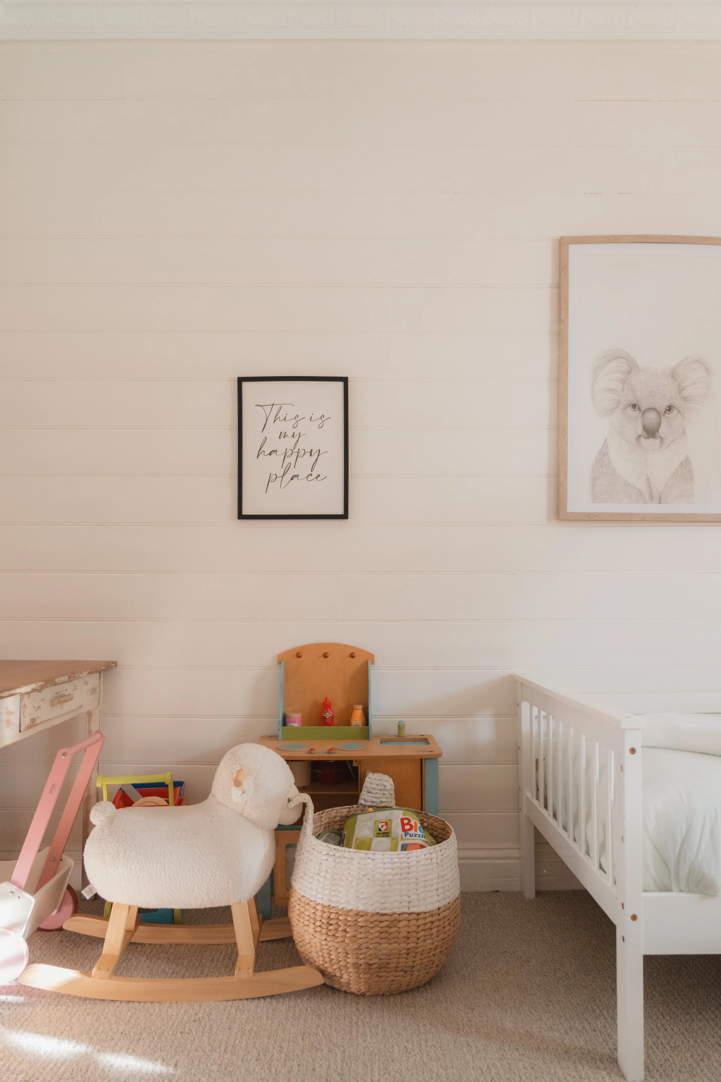 A corner of a child's nursery with light-colored horizontal wooden wall panels. Decor includes framed wall art, one with the phrase 'This is my happy place' and a drawing of a koala. Furniture includes a white crib, a wooden rocking chair with a plus