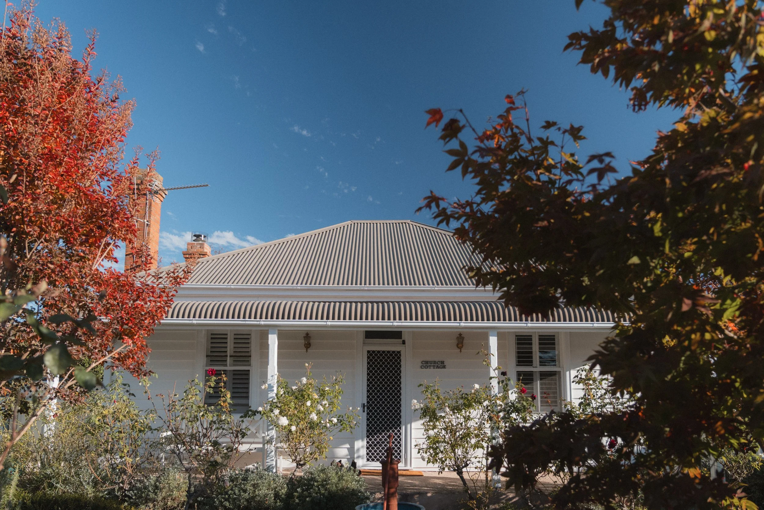 Front view of a house called 'Church Cottage' with a corrugated metal roof, surrounded by autumn trees with red, orange, and green foliage, under a clear blue sky.