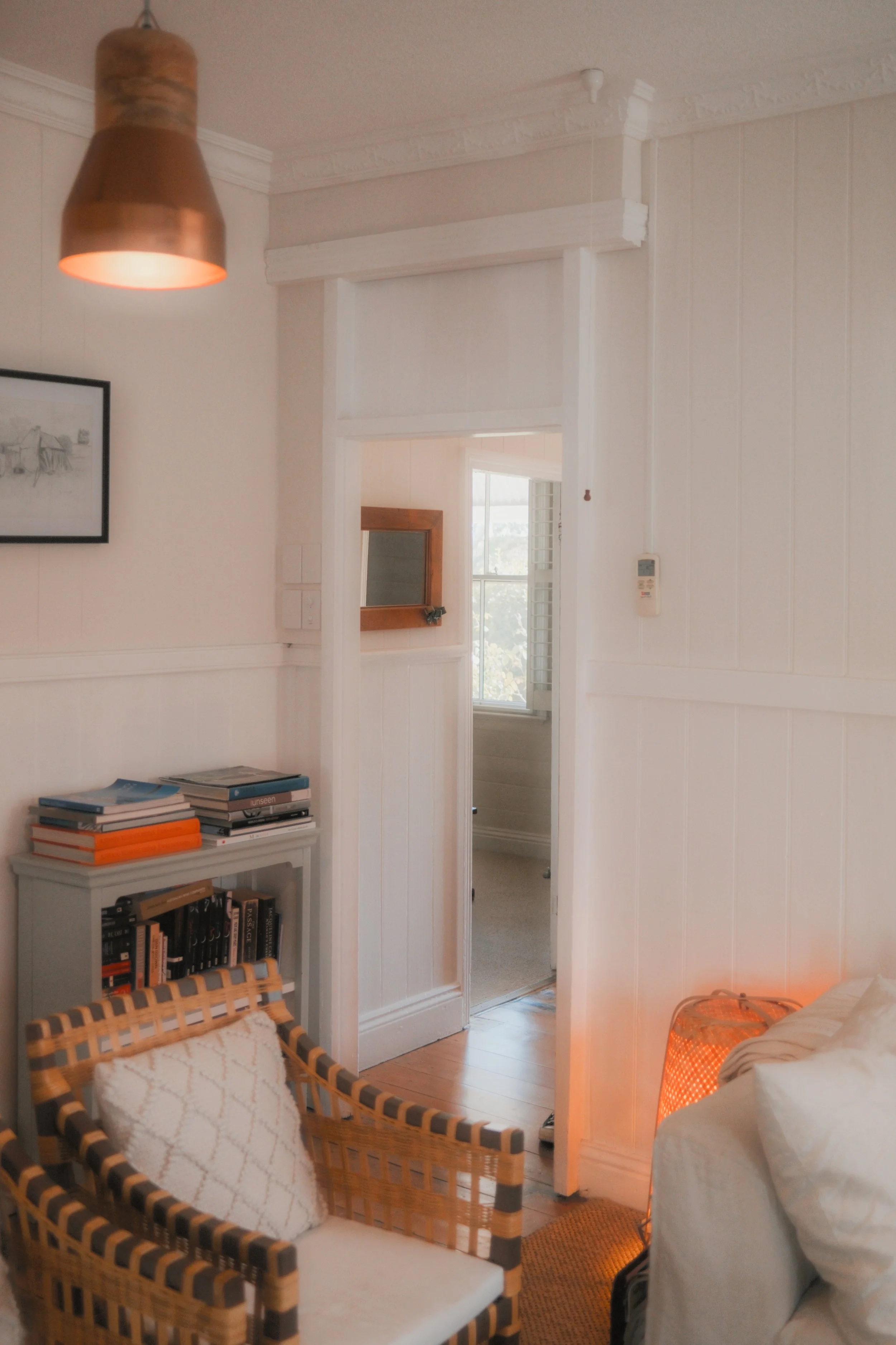 A cozy living room with a woven wicker chair with a white cushion and pillow, a small gray bookshelf with books, a framed black-and-white picture on the wall, a ceiling light, an orange lampshade on the floor, and a hallway leading to another room wi