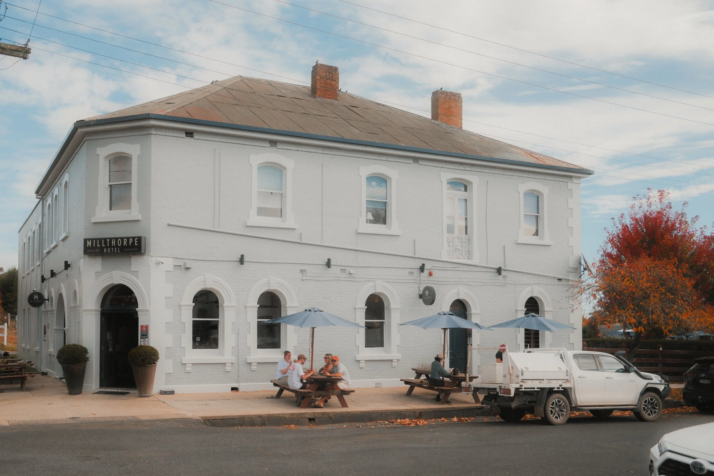 A two-story white brick building with a sign that reads "Millthorpe Hotel" on the corner. Outdoor seating with wooden picnic tables and umbrellas, and a few people sitting and conversing. Several cars parked in front, and a tree with red leaves on th
