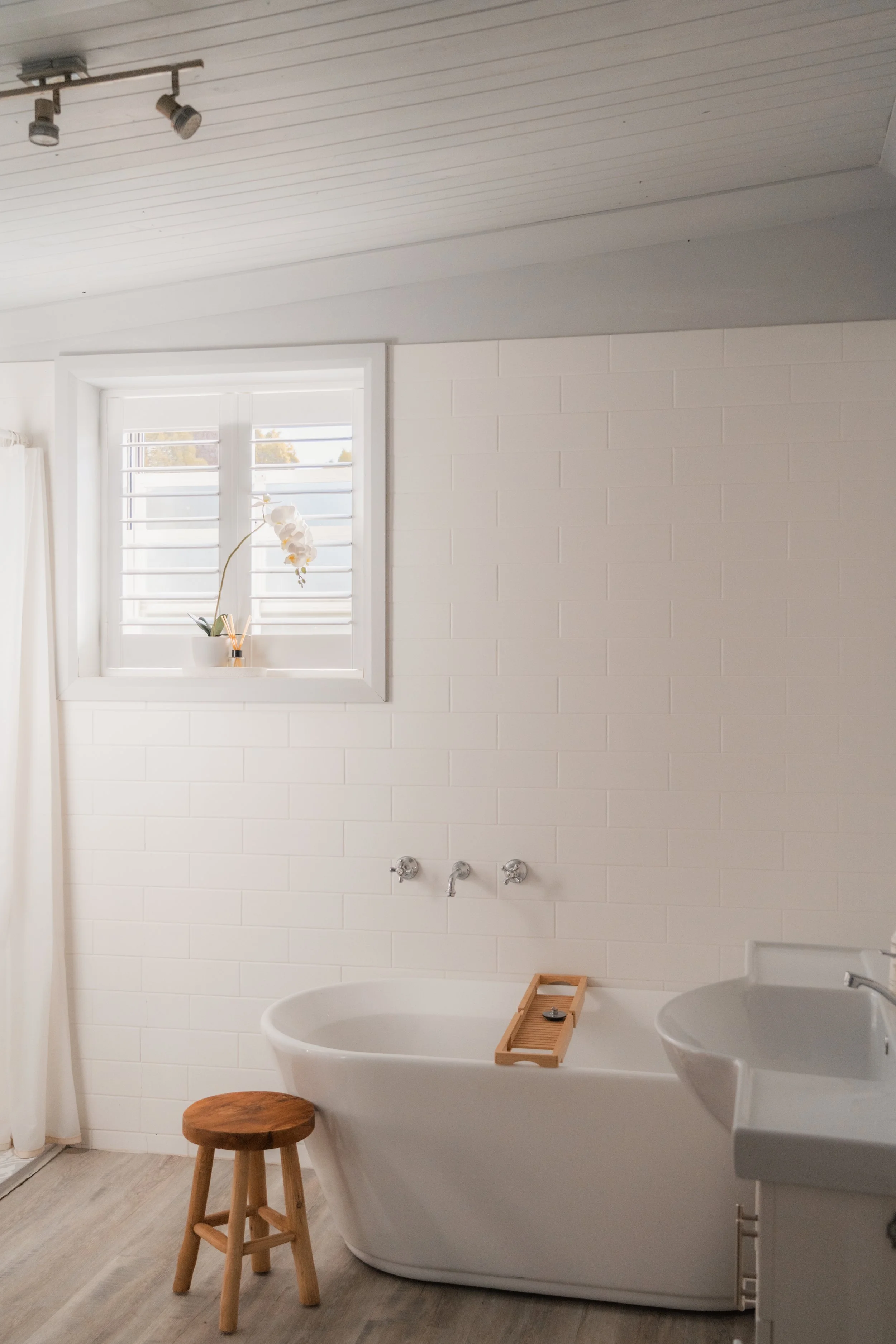 A minimalist bathroom with a white freestanding bathtub, a small wooden stool, a white sink, and a window with white shutters. There is a orchid plant on the windowsill and a wooden bath tray across the bathtub.