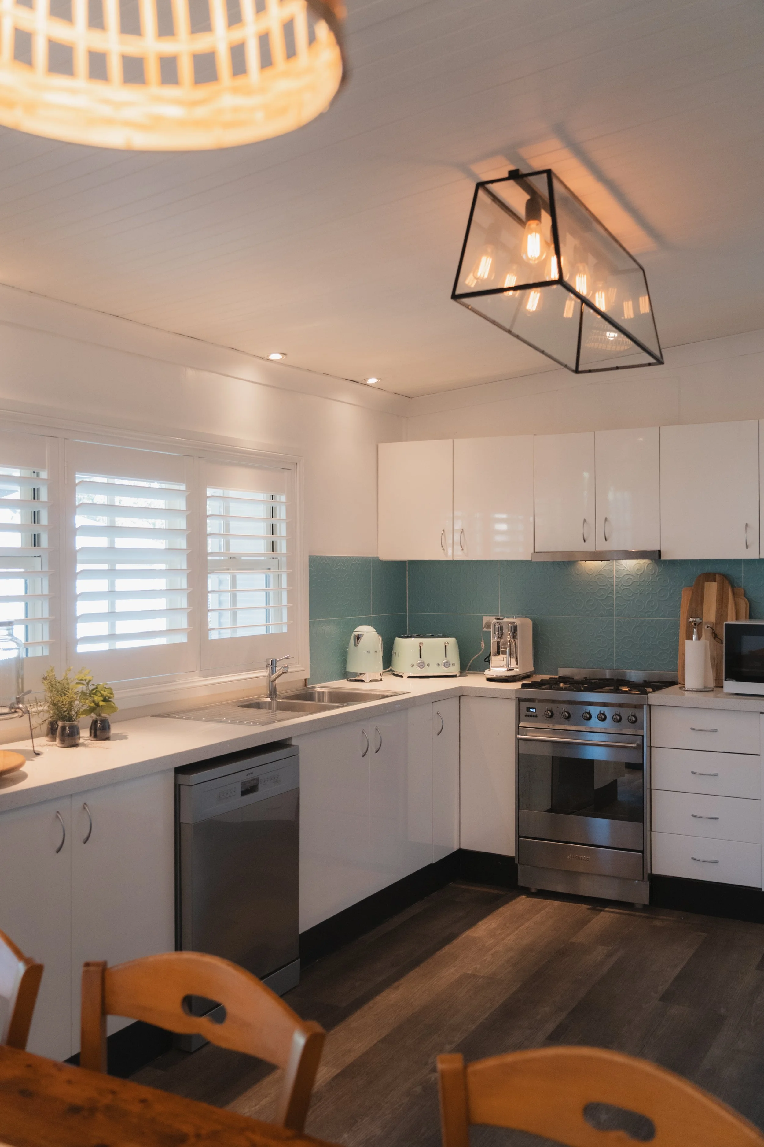 Modern kitchen with white cabinets, stainless steel stove, green textured backsplash, countertop appliances, window with white shutters, and wooden dining chairs.
