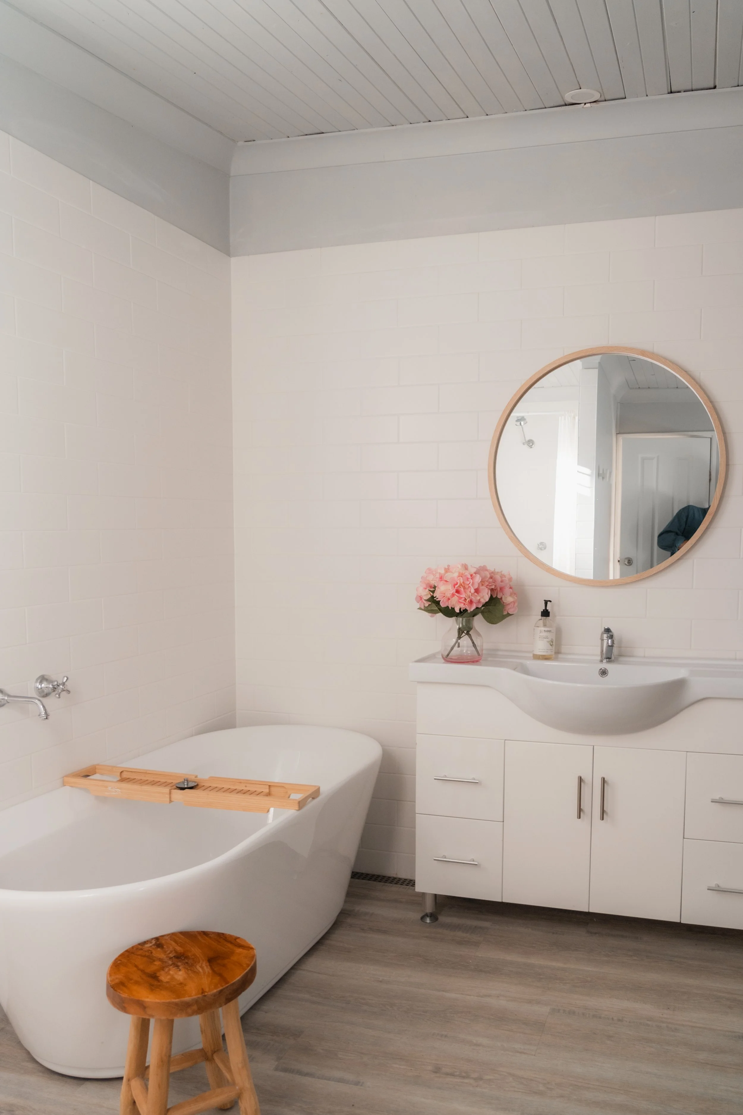 A modern bathroom featuring a white freestanding bathtub with a wooden bath tray, a white vanity with a small sink, a round mirror, a vase of pink flowers, and a stool, with light gray wood flooring and simple white walls.