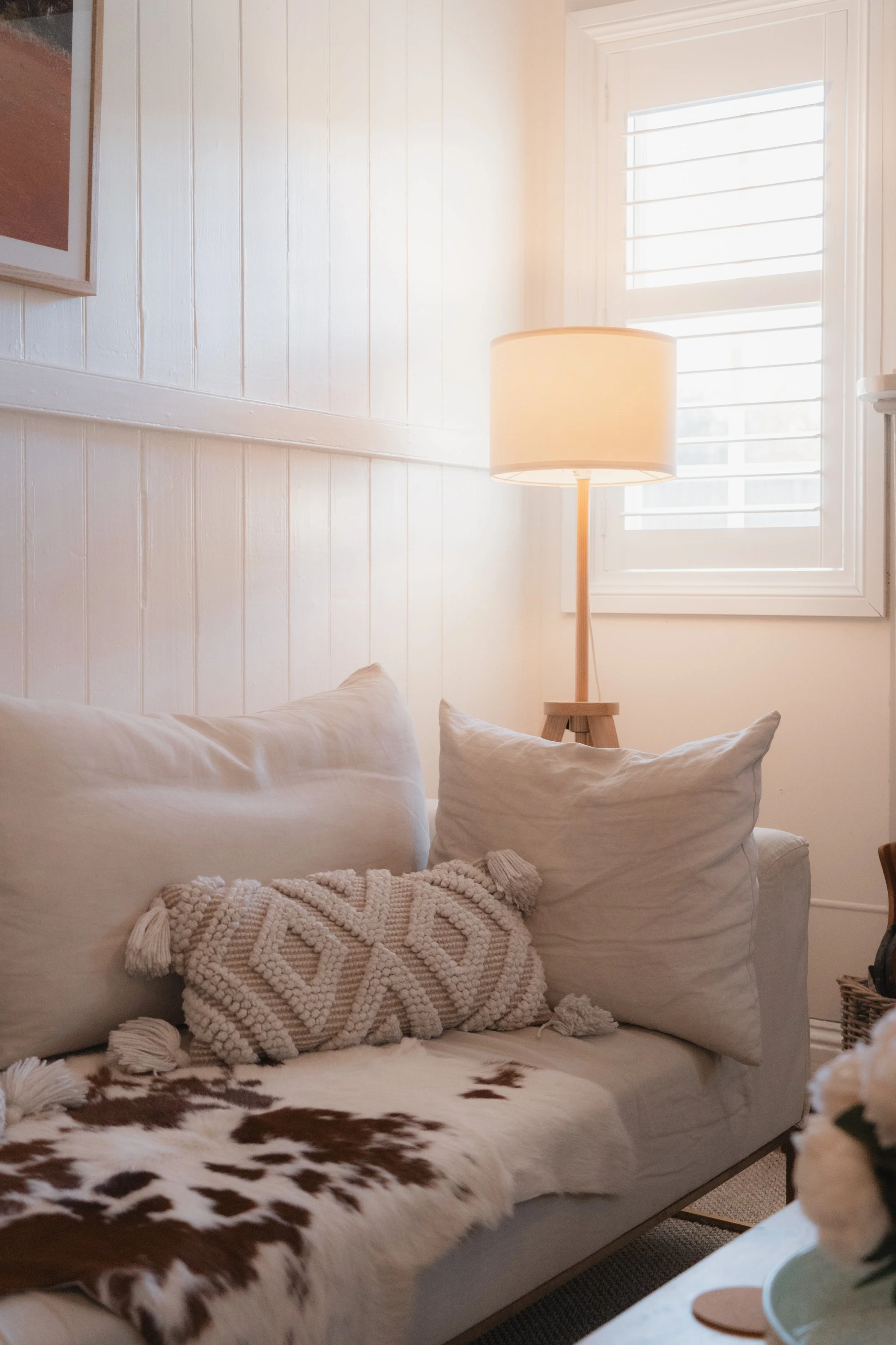 A cozy bedroom corner with a white bed, cowhide blanket, decorative pillows, a wooden bedside lamp, and a window with white shutters.