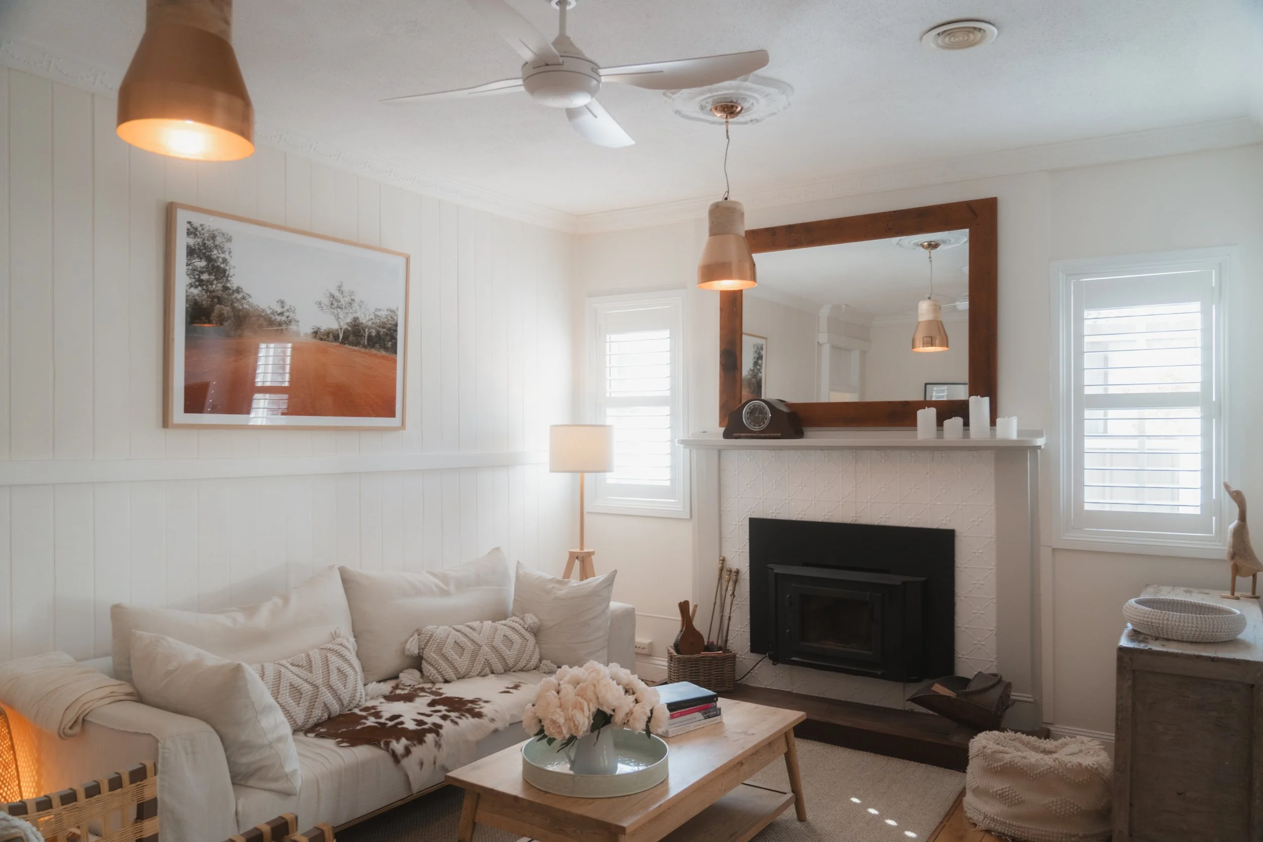Cosy white living room at Church Cottage with a cream sofa, timber coffee table, fireplace, large wall mirror, pendant lights and soft natural light through shuttered windows.