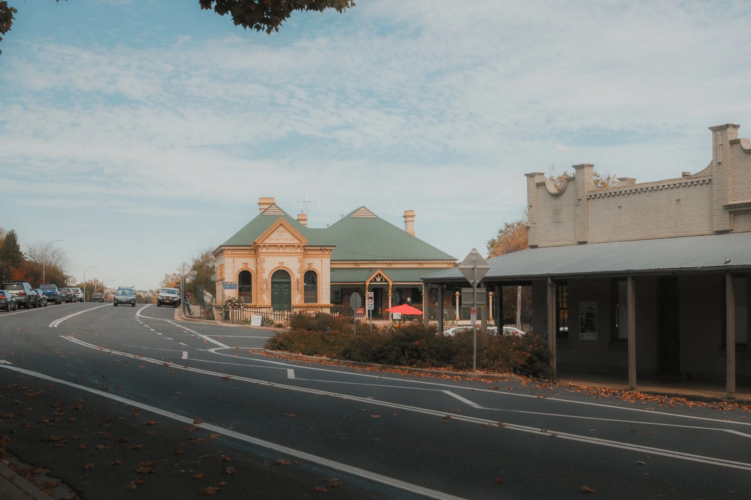 A small town street scene with a historic building having a green roof and ornate architecture, a row of parked cars along the street, trees with autumn leaves, and a church or similar structure with a white brick facade and decorative battlements. T