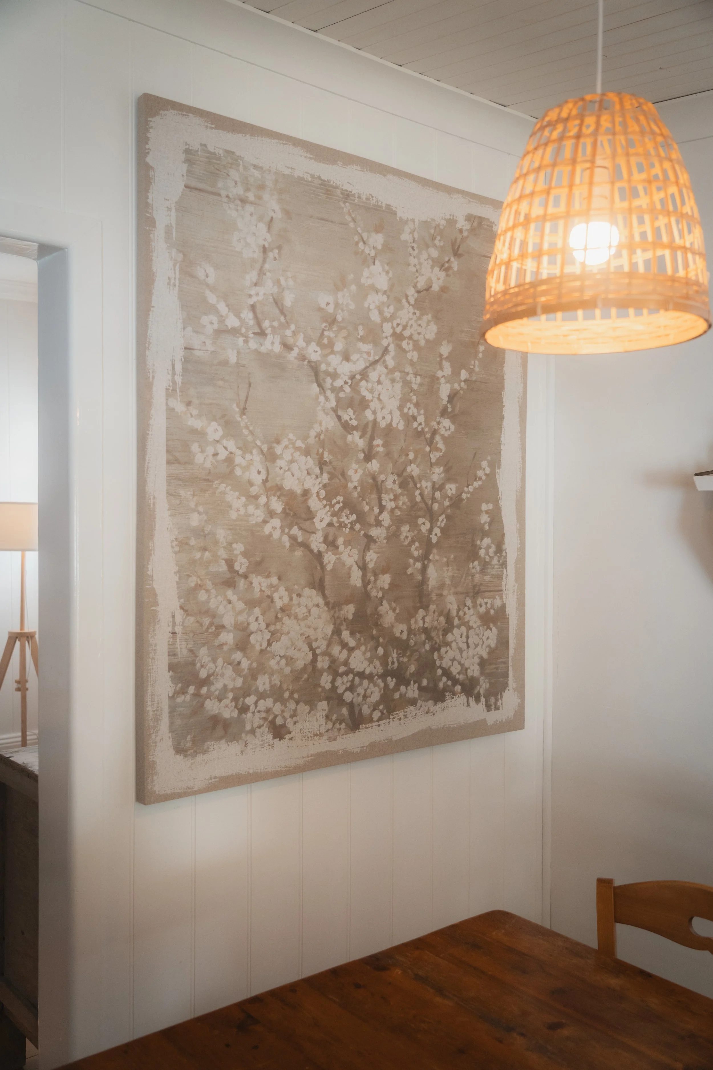 A beige and white floral Japanese cherry blossom painting hanging on a white-paneled wall above a wooden chair and table, with a rattan pendant light illuminating the room.