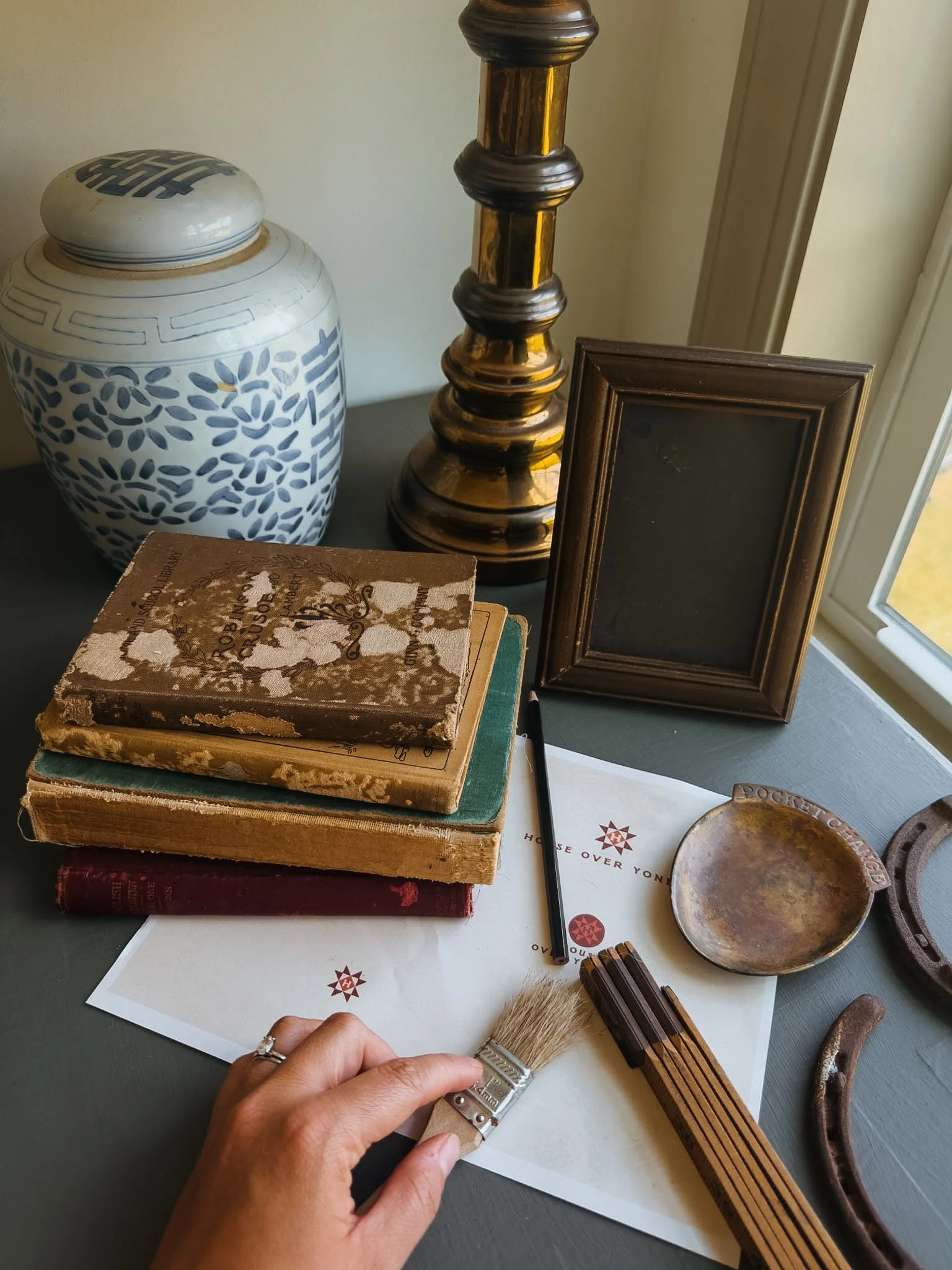 A person touching a small paintbrush laid over a piece of paper on a table. The table also has a stack of vintage books, a decorative vase, a lamp post, a small frame, a round metal dish, and some other small objects.