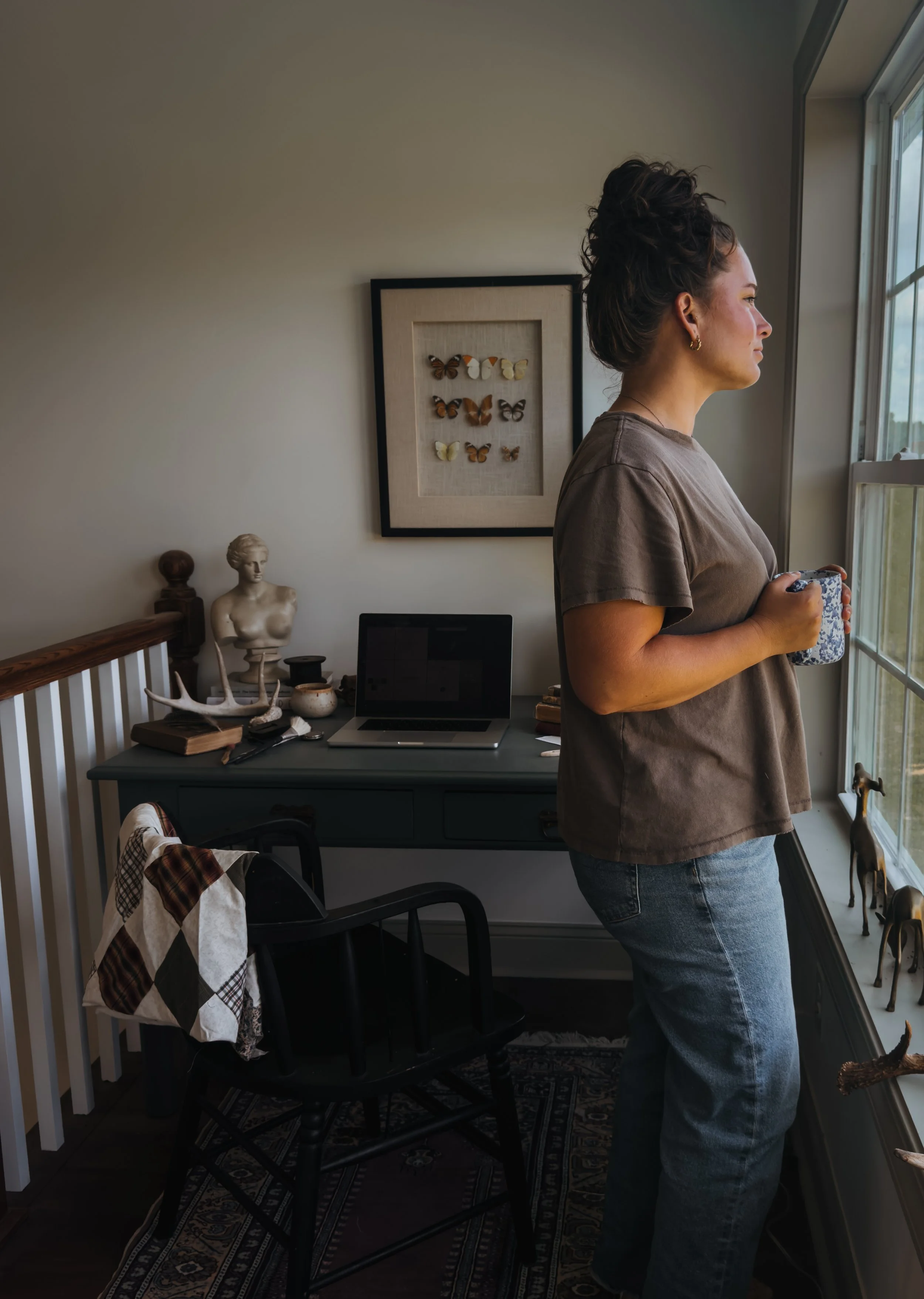A woman with curly hair in a messy bun wearing a gray T-shirt and jeans, looking out a window while holding a mug, inside a room with various decorative items and artwork.