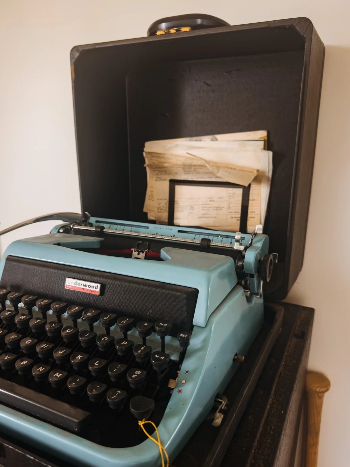 A vintage blue Underwood typewriter with a sheet of paper inserted, placed on a wooden surface with a black case open behind it.