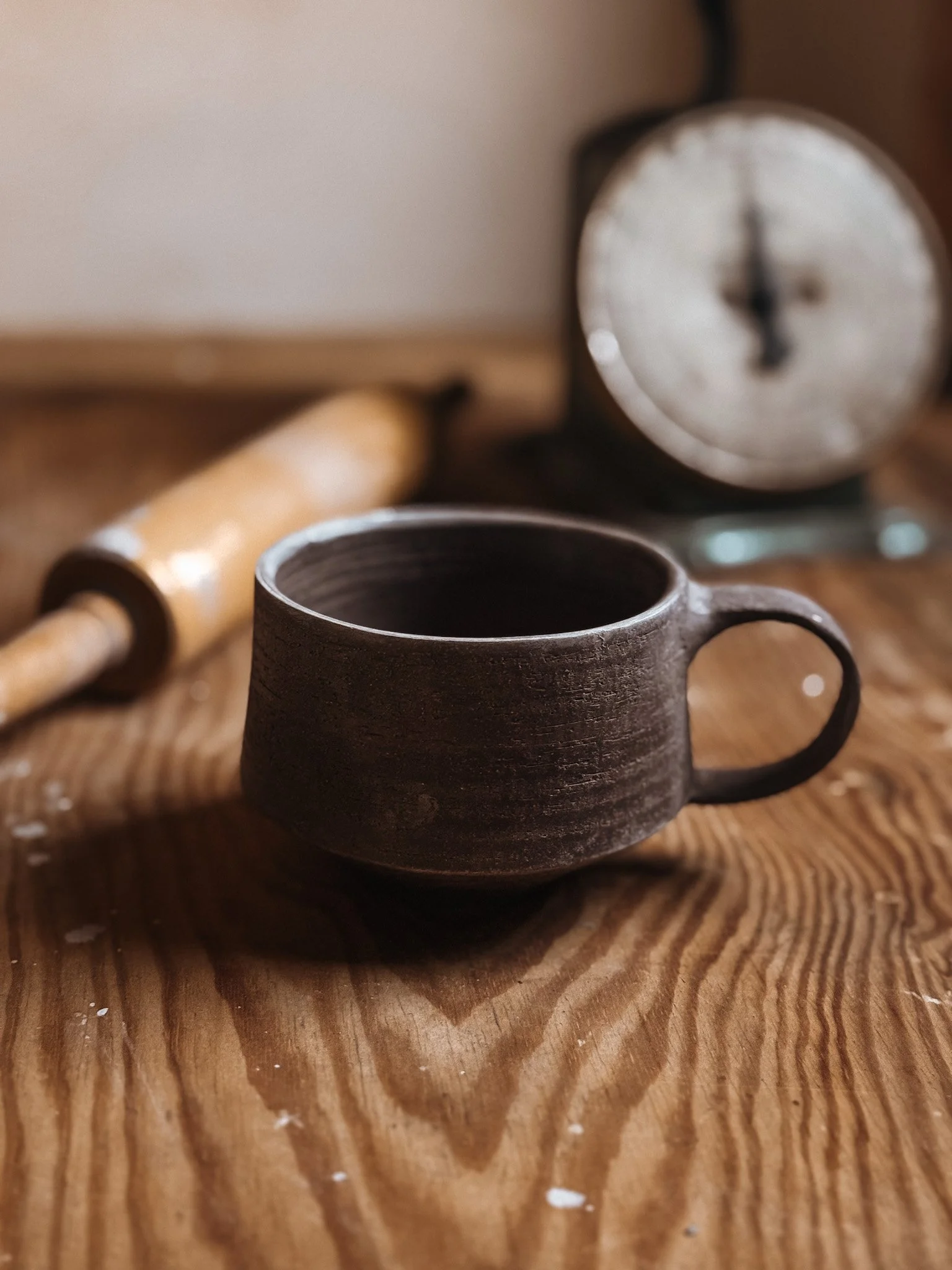 A dark brown ceramic mug with a handle on a wooden surface, with a vintage clock in the background and a rolling pin nearby.