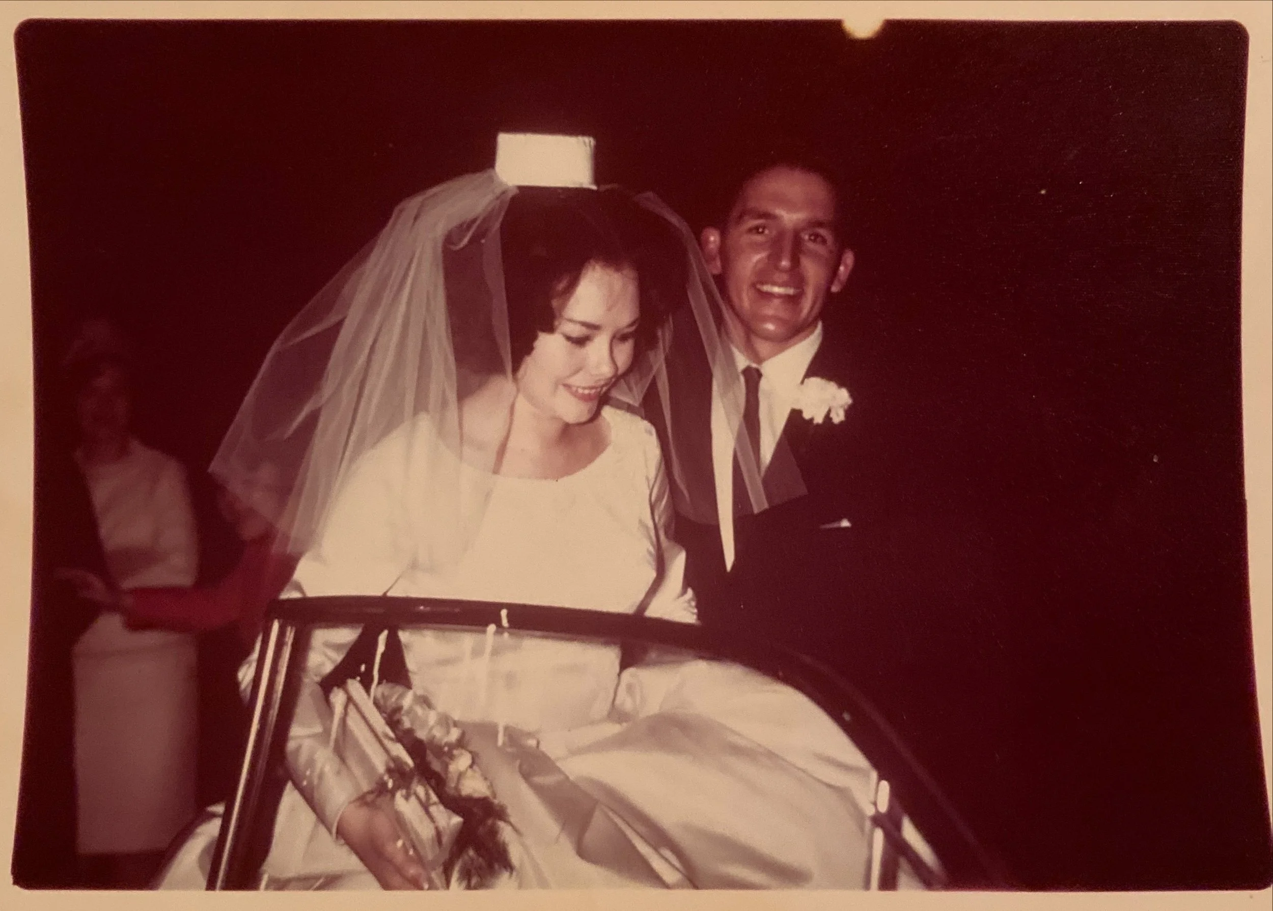 A black-and-white wedding photo of a smiling bride and groom sitting in a vintage car, with the bride wearing a veil and holding flowers.