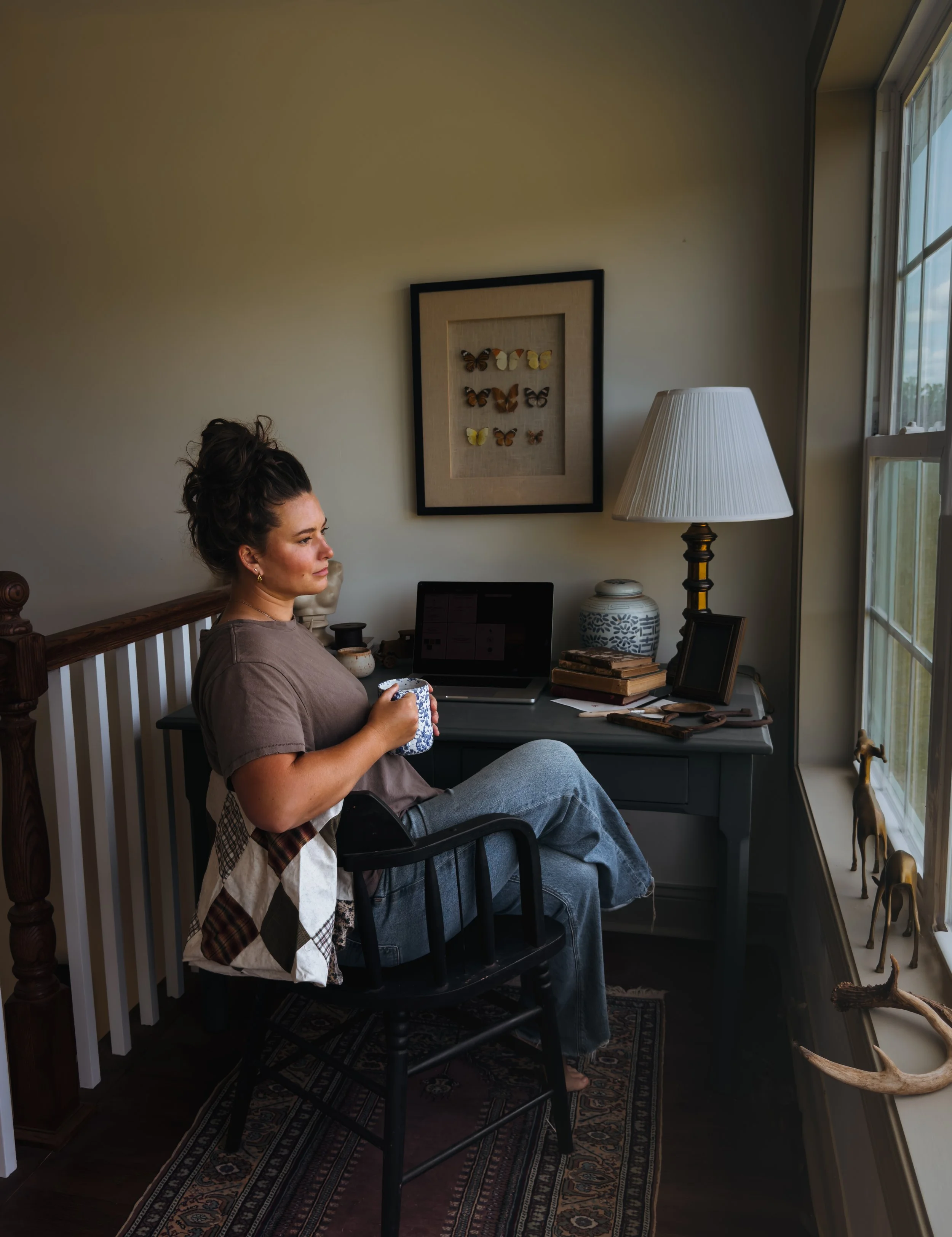 A woman sitting in a black wooden chair in a cozy home office, holding a mug, with a window on her right and a framed butterfly collection on the wall behind her.