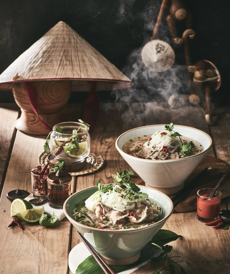 Deux bols de soupe vietnamienne avec des herbes fraîches, une lime, et des sauces, sur une table en bois avec un décor traditionnel vietnamien.