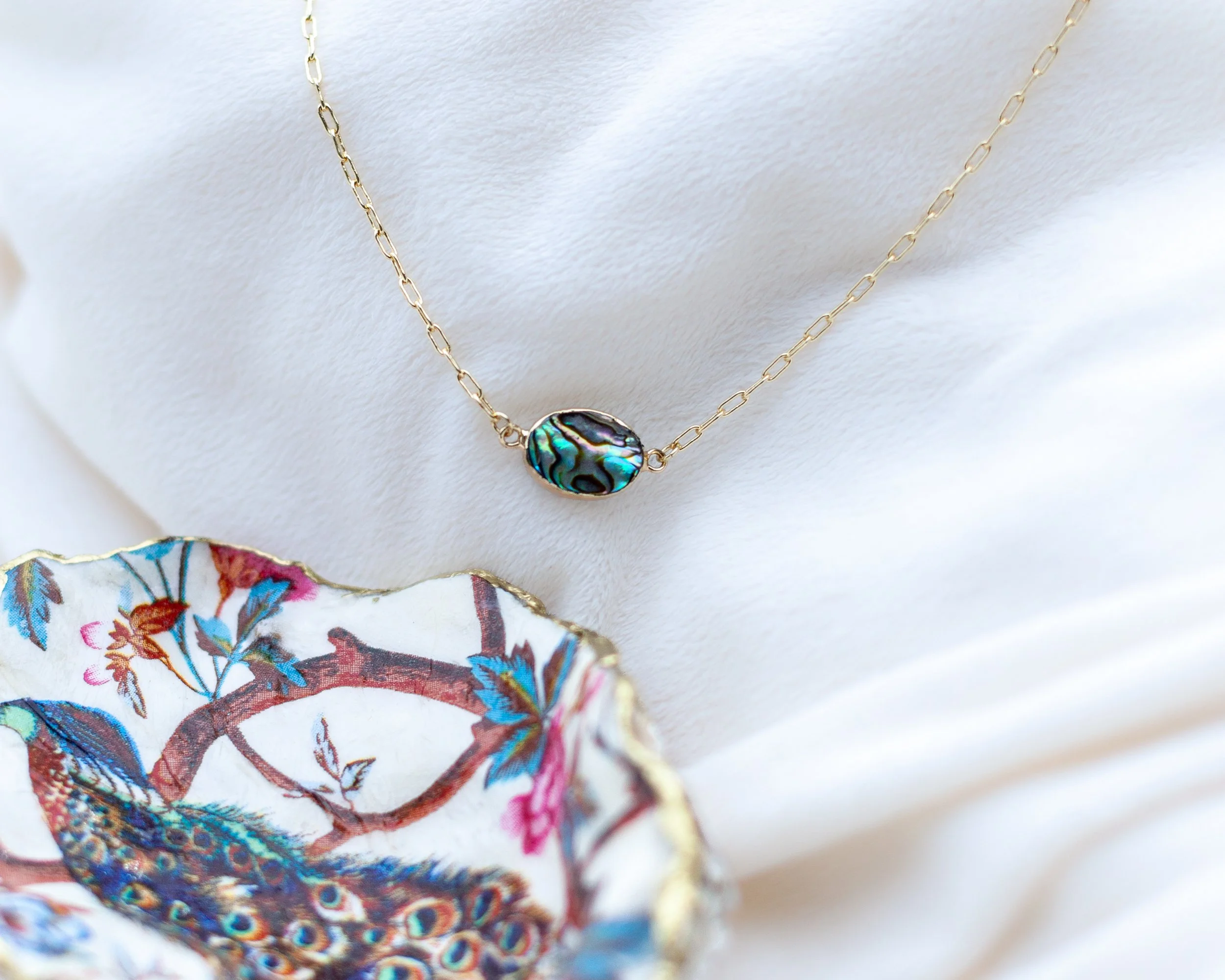 A gold necklace with a large abalone shell pendant hanging on a white fabric background.