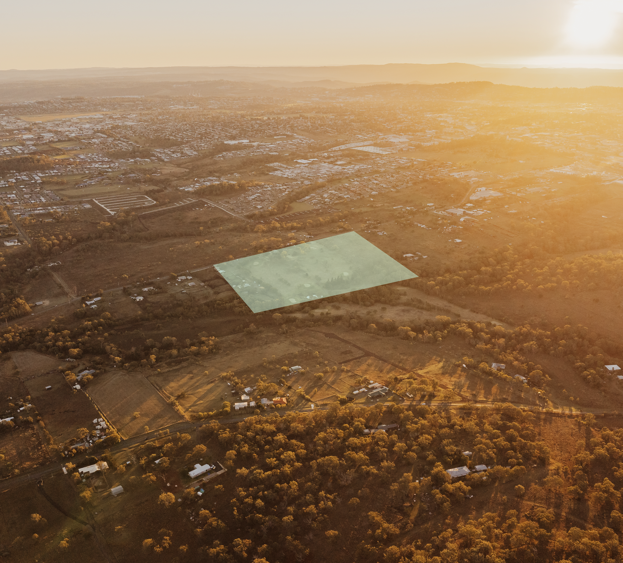 Aerial view of a rural area during sunset, showing fields, roads, scattered houses, and a small green field, with a haze and mountain range in the background.