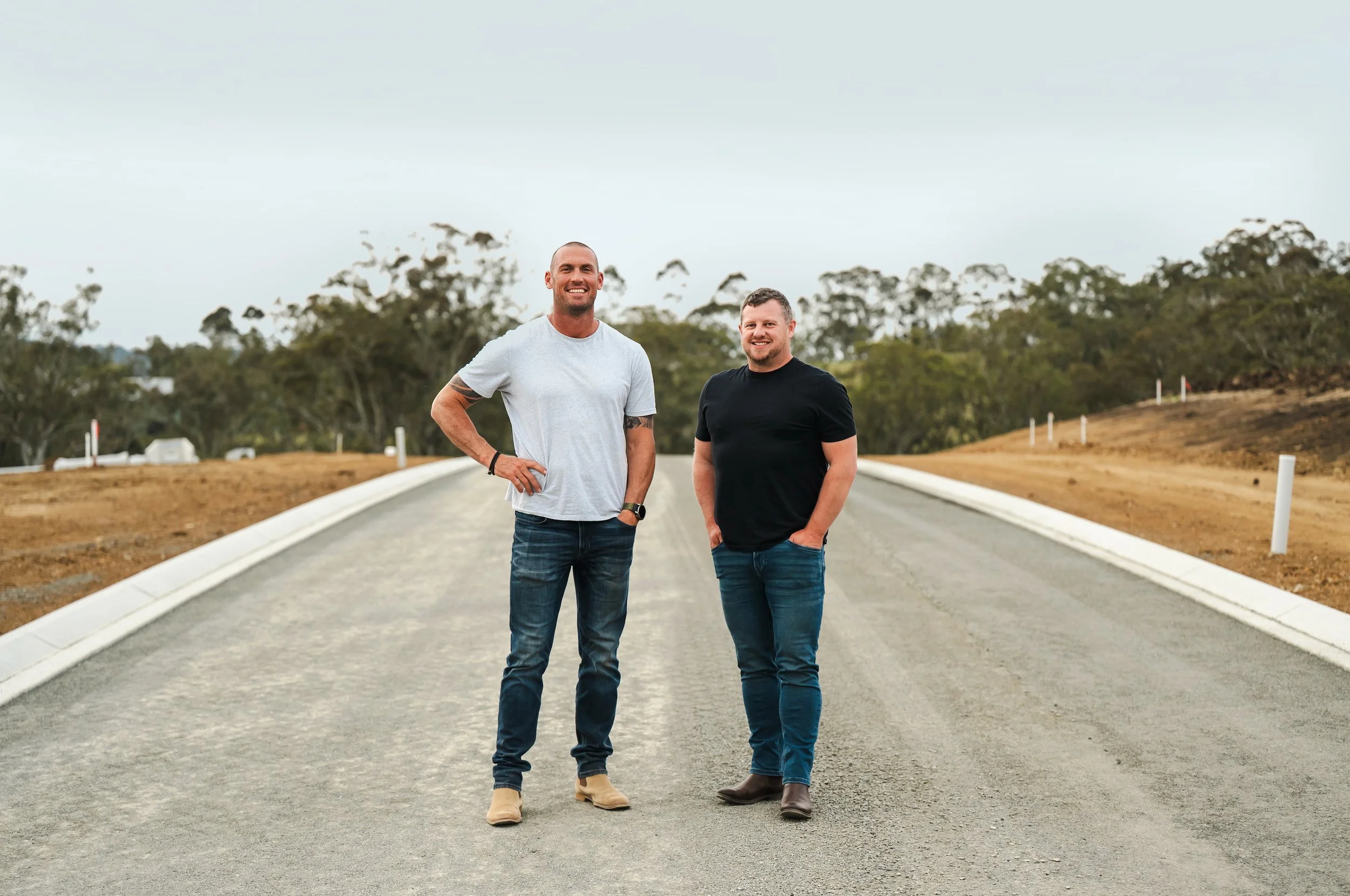 Two men standing on an empty road with trees in the background, smiling at the camera, one in a white t-shirt and the other in a black t-shirt.
