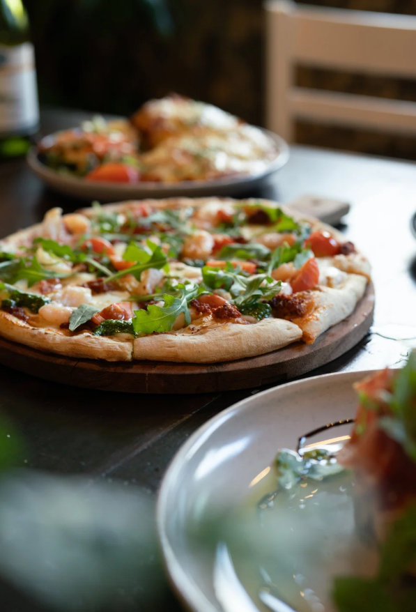 A pizza with cheese, tomatoes, and fresh basil on a wooden serving board, with other dishes on a dark table.