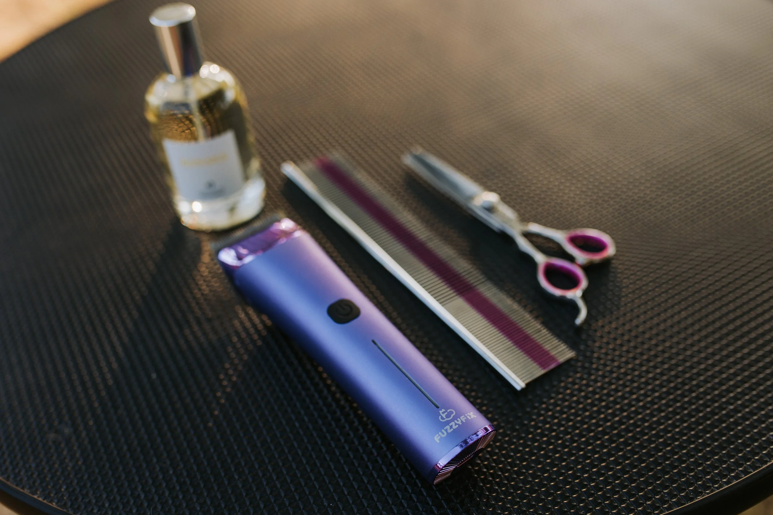 Hair cutting tools including a comb, scissors with pink handles, a purple electric hair clipper, a bottle of hair product, and a hair straightener on a dark textured surface.
