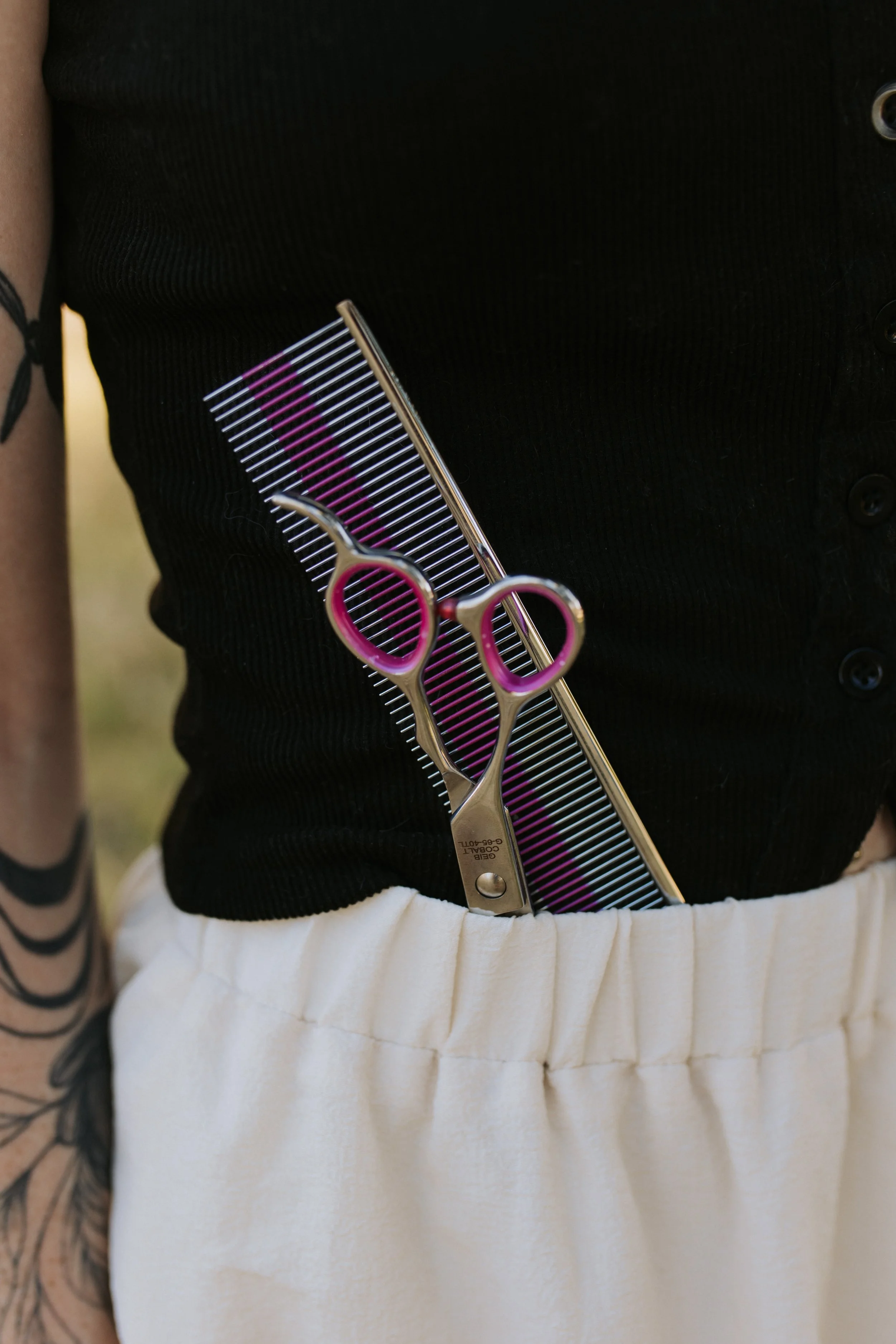 Person wearing a black shirt and white pants, holding a pair of hair thinning scissors and a black and purple comb.
