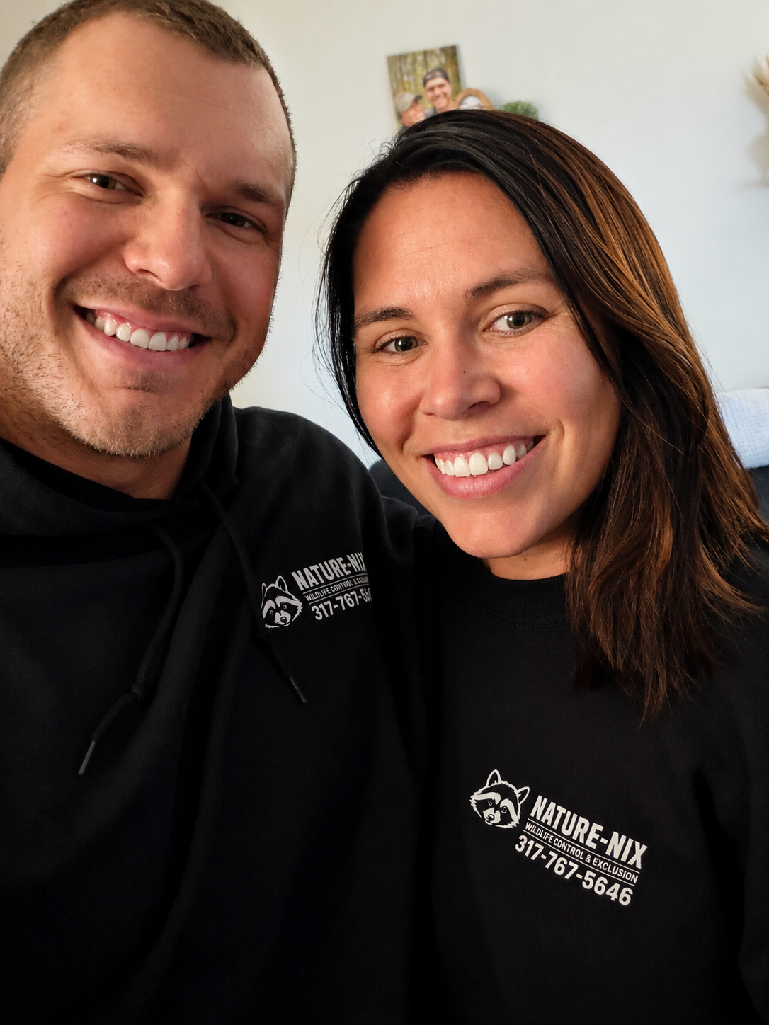 A smiling man and woman taking a close-up selfie indoors, both wearing black shirts with white text and a raccoon logo for NATURE-NIX, a wildlife control and exclusion company.