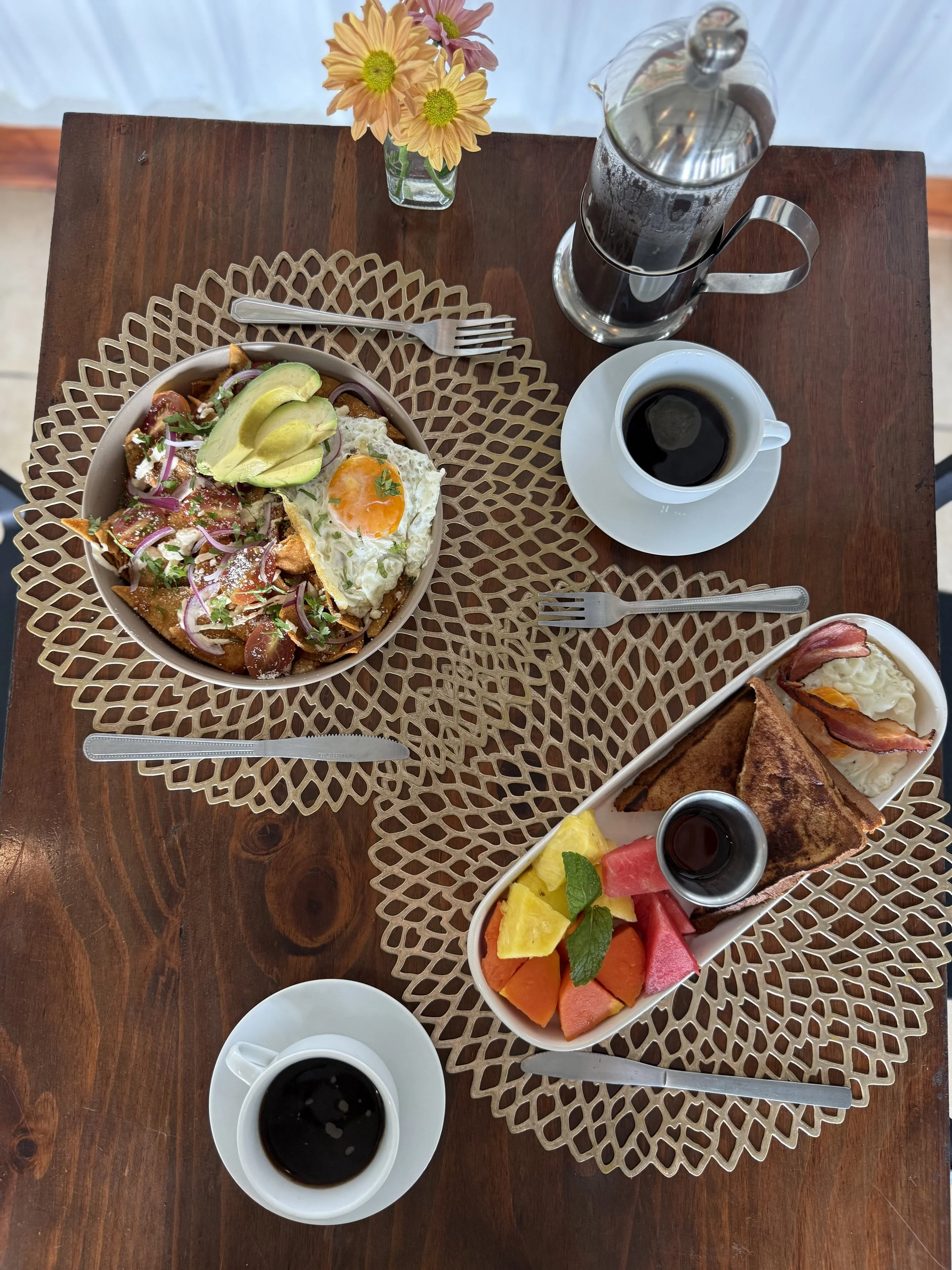 Desayuno con huevos fritos, aguacate, tomate, cebolla, papas, pan tostado, frutas variadas, café y agua con hielo sobre una mesa de madera. También hay un florero con flores amarillas y rosas.
