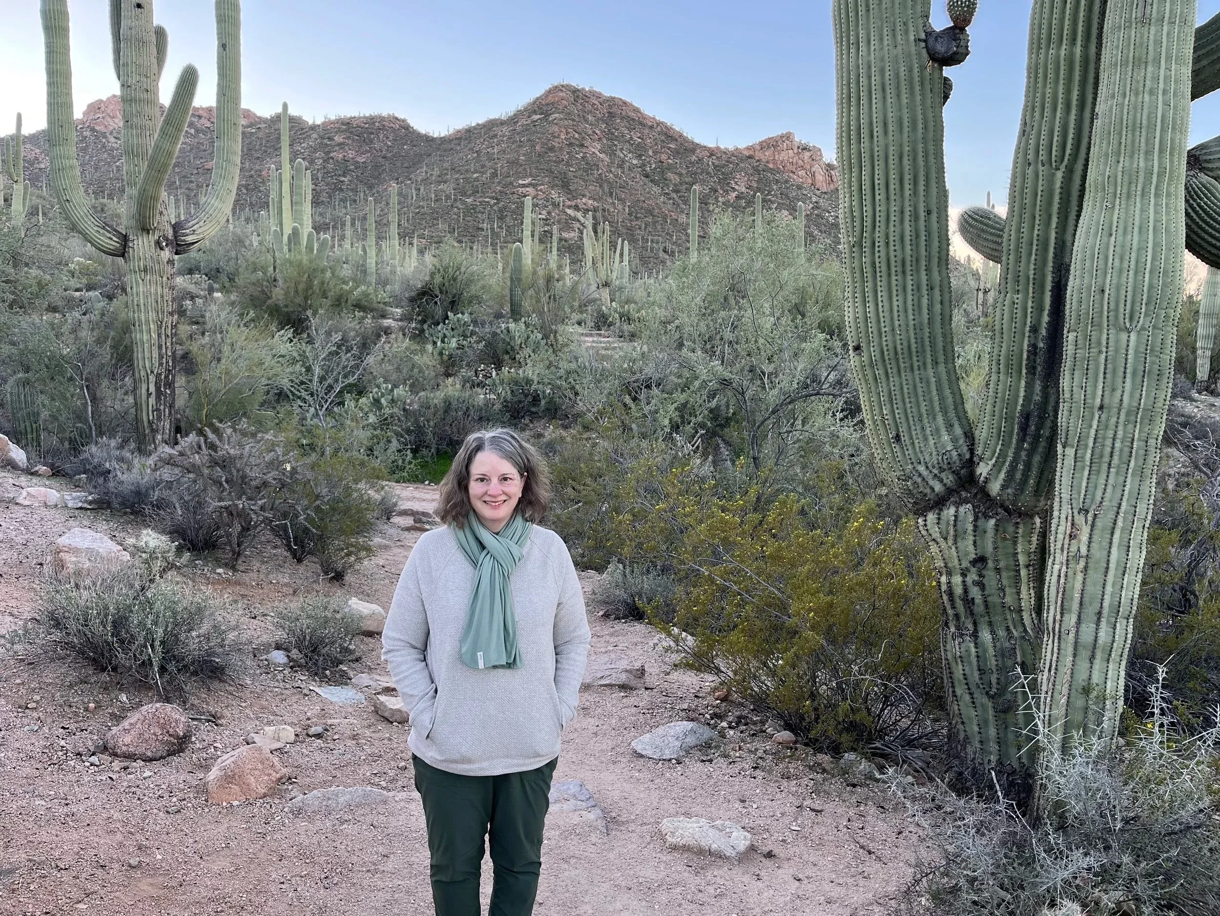 Janice Maust Hedrick standing on a desert trail among giant saguaro cacti in Arizona.