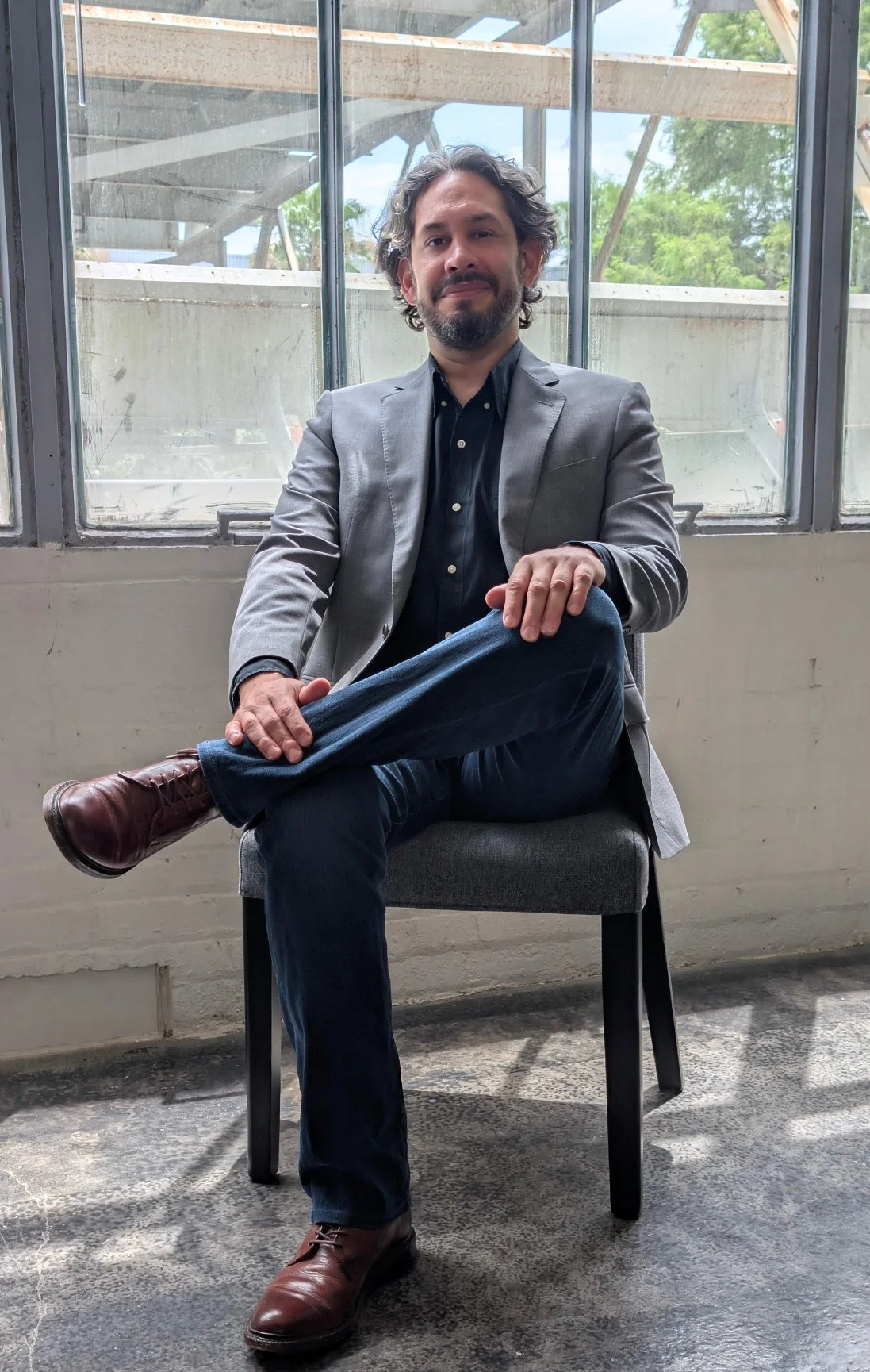 A man with curly dark hair and a beard sitting on a chair in front of large windows, wearing a gray blazer, black shirt, jeans, and brown leather shoes, crossing his legs with his right ankle on his left knee.