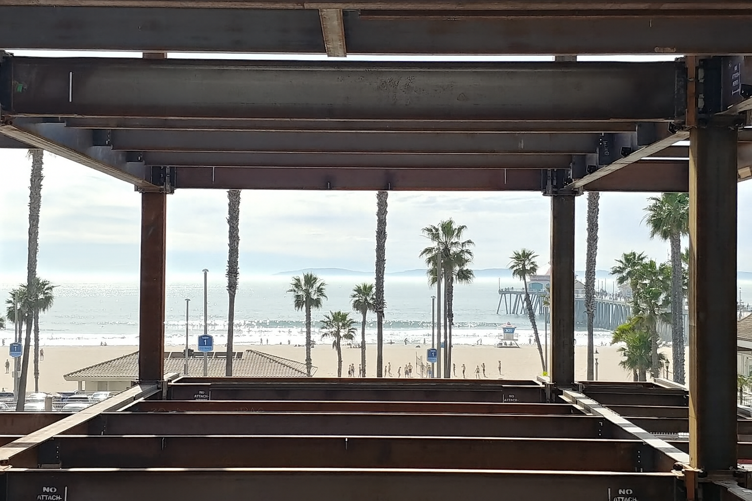 View of a beach and ocean through the framework of a building under construction, with palm trees and a pier in the distance.
