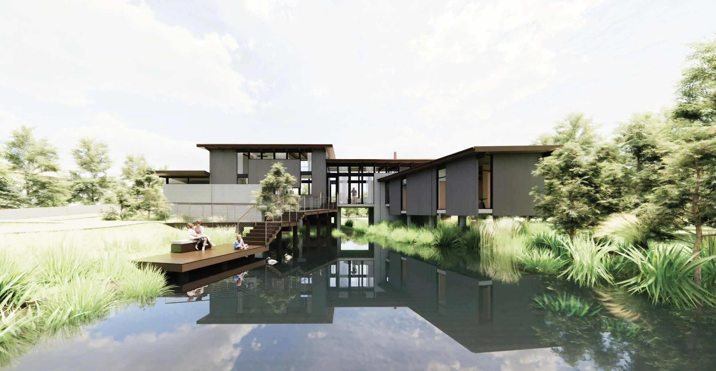 Modern house with large glass windows and gray exterior, surrounded by trees and greenery, with a pond reflecting the house and sky, and people sitting on a deck by the pond.