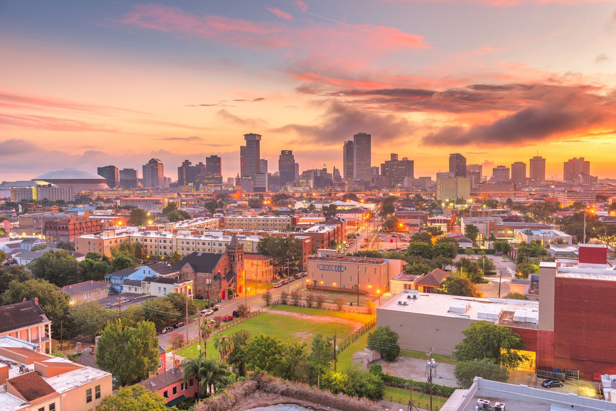 A city skyline at sunset with tall buildings, a domed sports arena, residential houses, and a church with a steeple in the foreground.