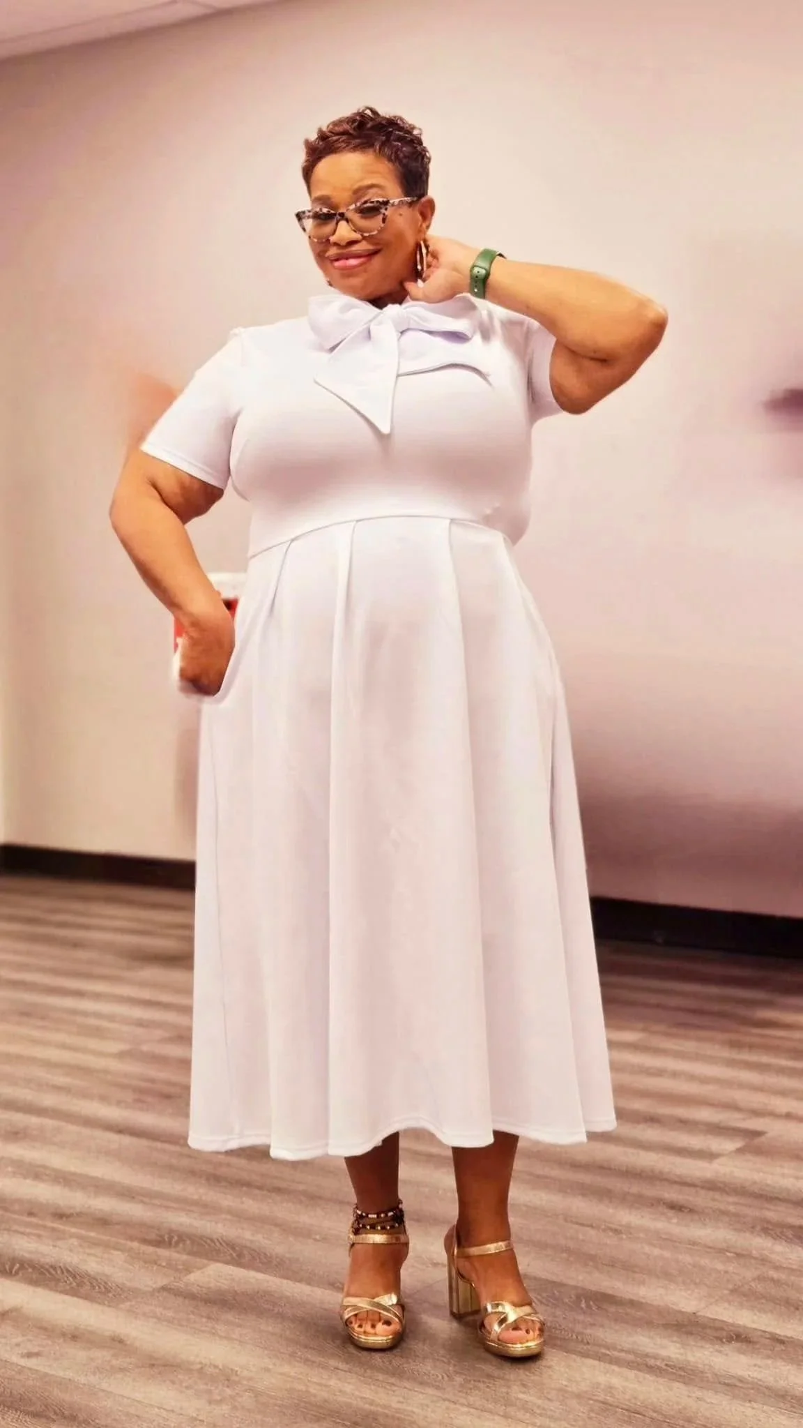 A woman in a white dress with a bow at the collar, wearing glasses, gold high heels, and a green wristband, smiling and posing indoors.