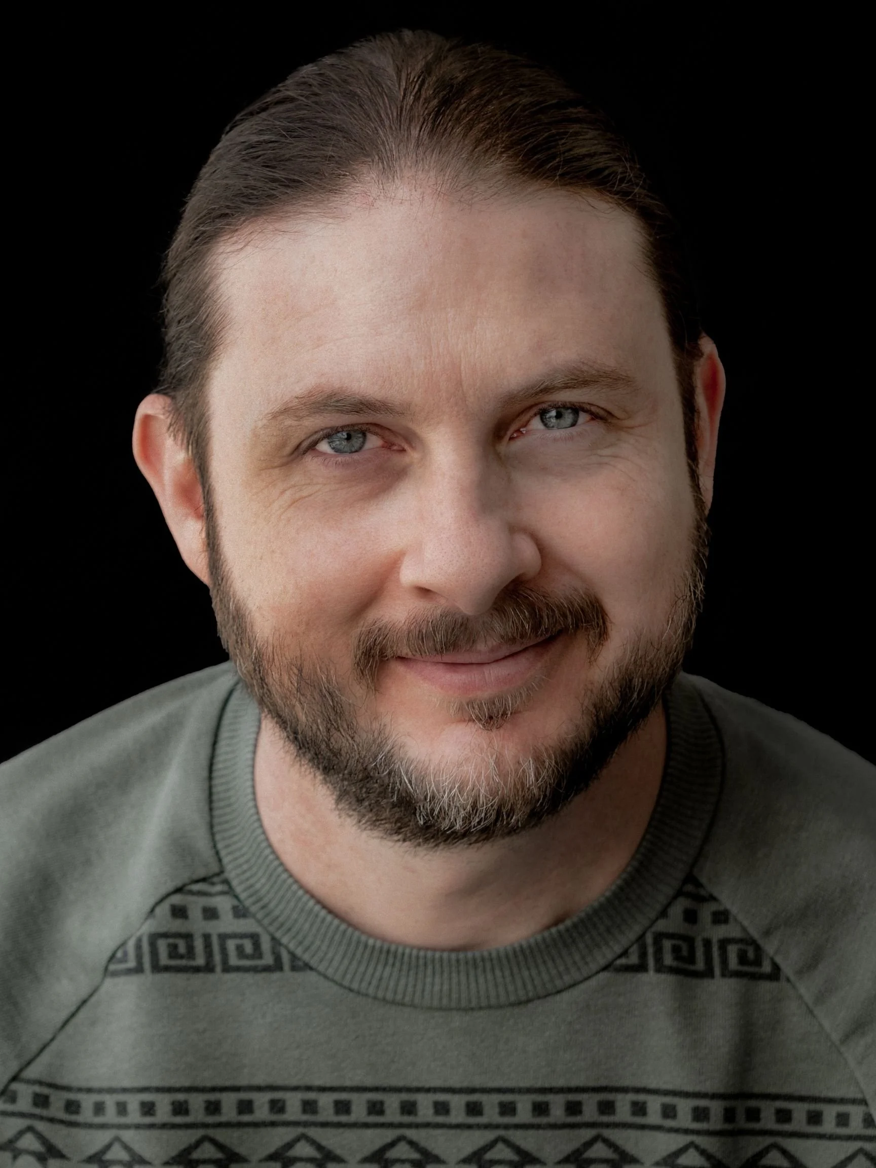 Close-up of a smiling man with blue eyes, brown hair, and a beard, wearing a gray patterned sweater against a black background.