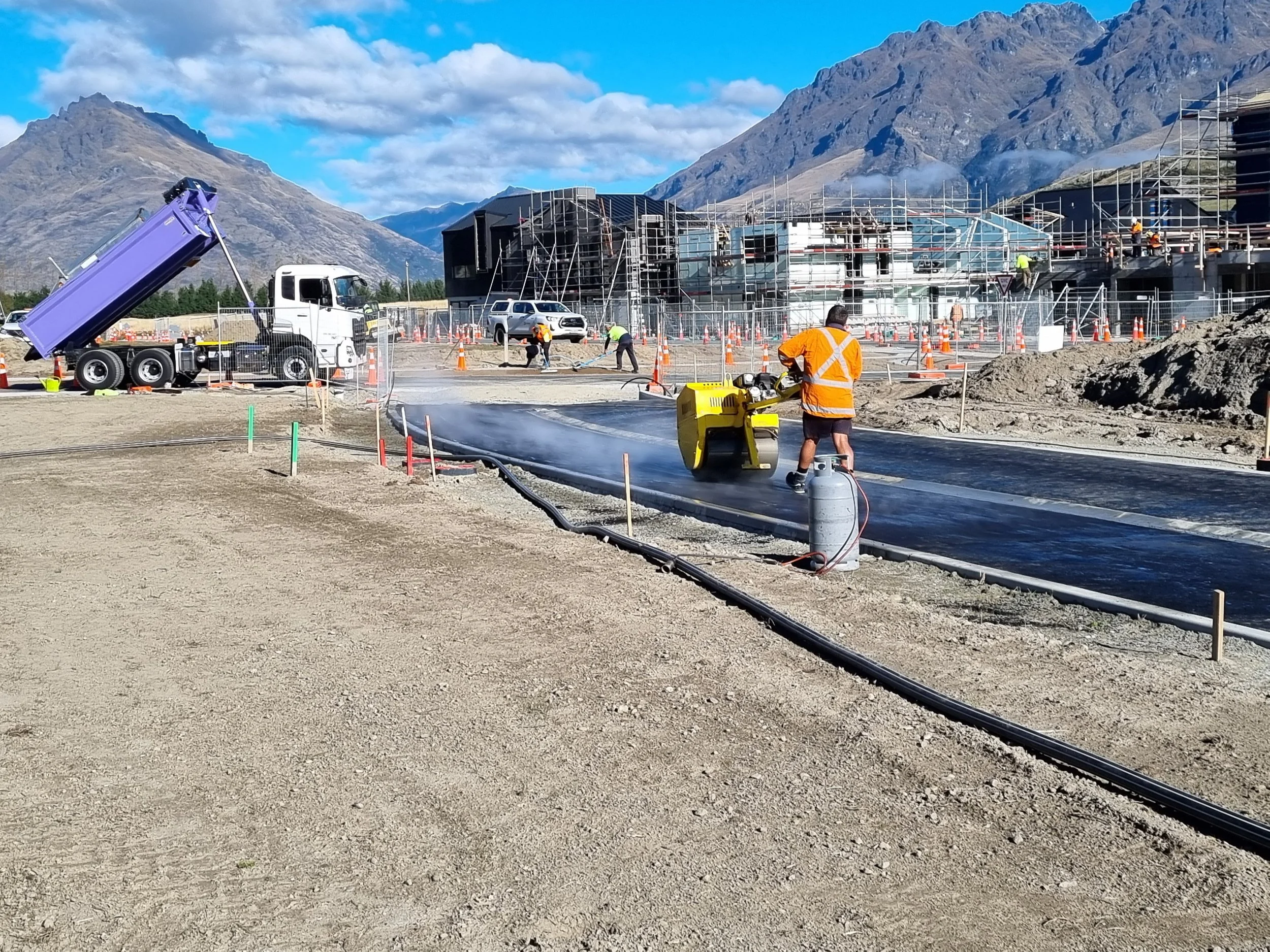 Construction site with workers laying concrete, scaffolding around buildings, a dump truck pouring concrete, and mountains in the background.