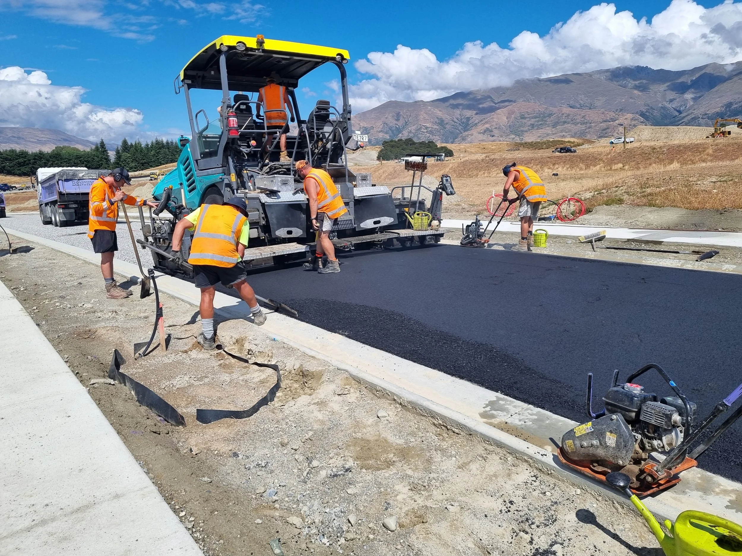 Construction workers paving a road with asphalt in a mountainous area under a partly cloudy sky.