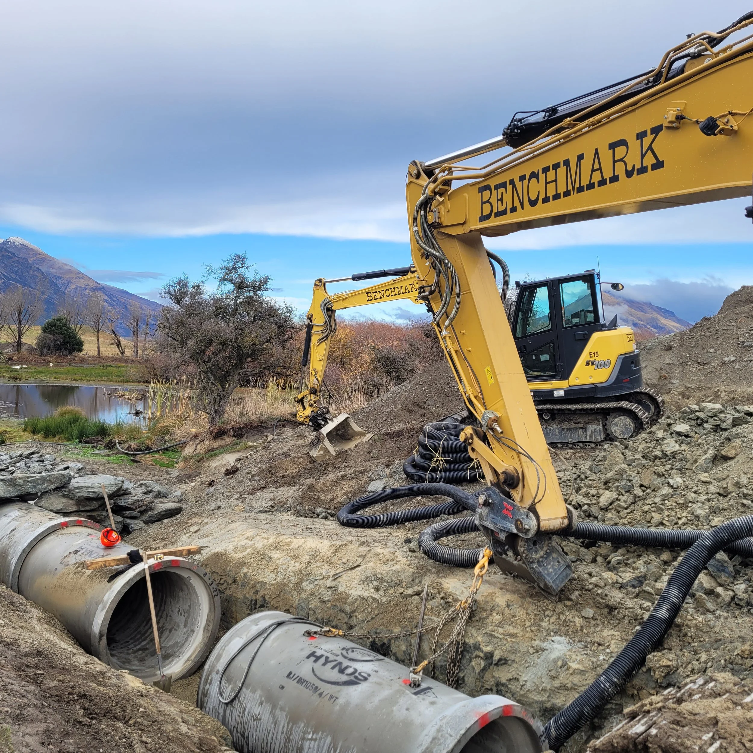Construction excavator digging near a large pipeline in a mountainous area with trees and cloudy sky for drainage