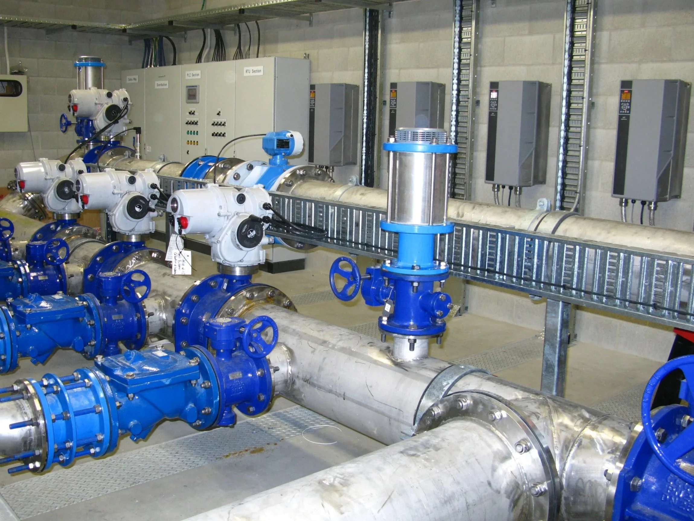 Industrial underground piping system with blue valves and metal pipes, and control panels mounted on a concrete wall in a utility room.