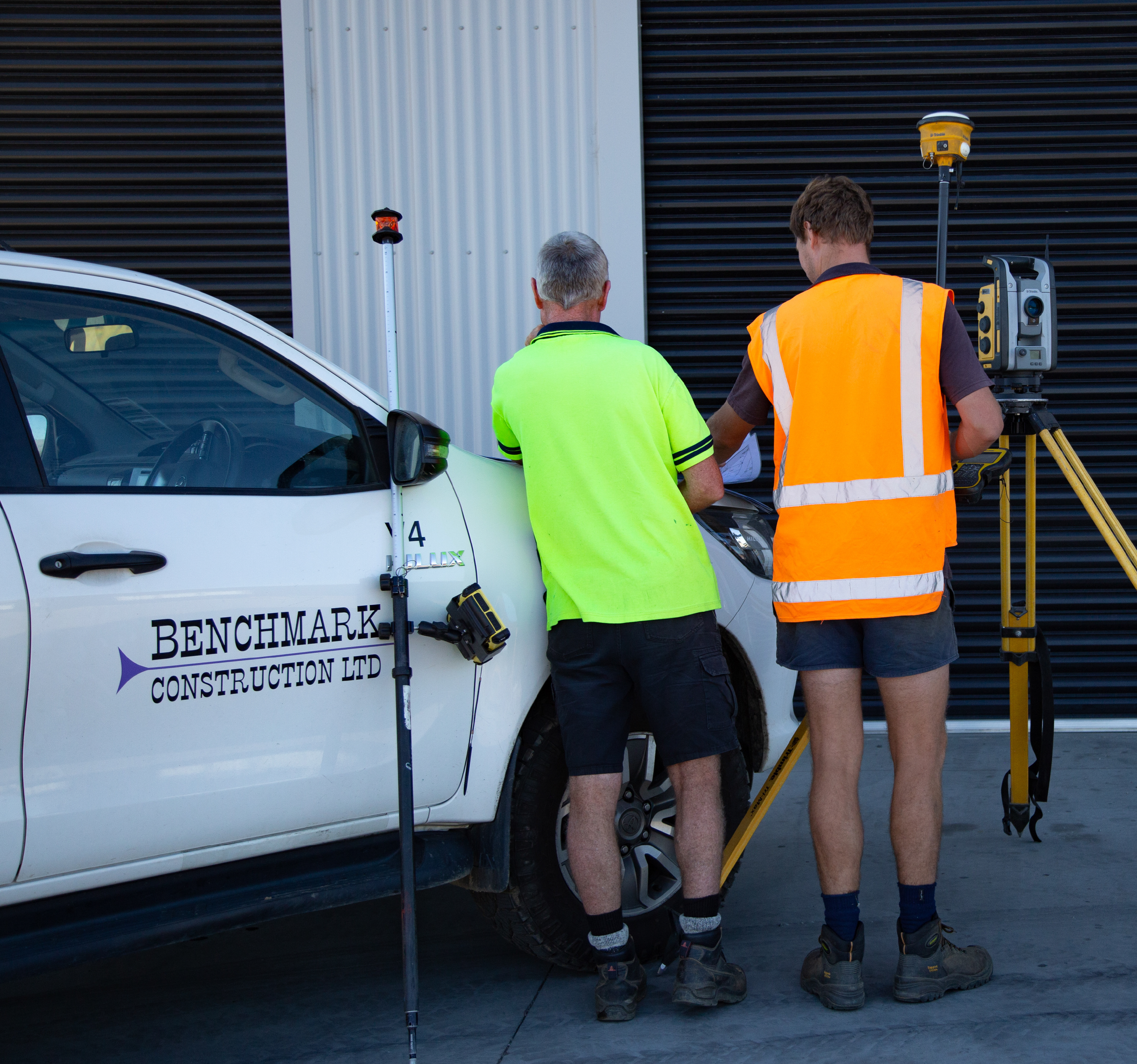 Two men in safety vests and shorts working on a construction site, using civil construction surveying equipment in a carpark with a partially visible door and a company logo that reads 'Benchmark Construction Ltd'.