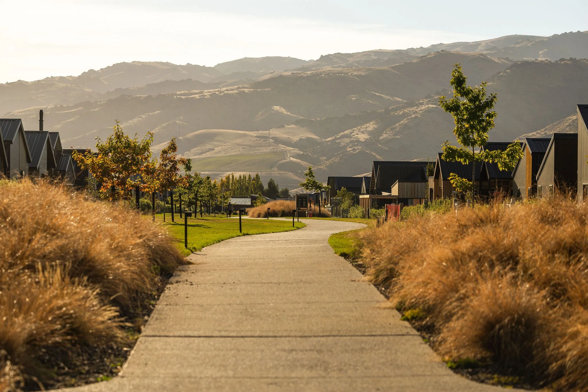 A paved walkway in a park with tall grasses on both sides, showcasing Asphalt surfacing by Benchmark Construction with hills in the background under a lightly cloudy sky in Cromwell, Central Otago.