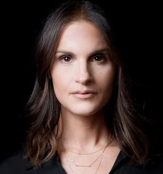 A portrait of a woman with long, wavy brown hair wearing a black top and layered gold necklaces, against a black background.