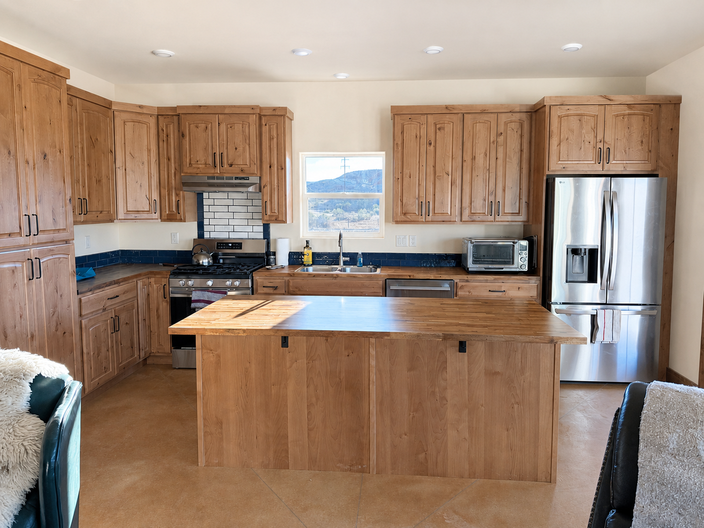 A kitchen with natural wood cabinets, a central wooden butcher block island, a stainless steel refrigerator, a stove, a microwave, and a small window with mountains in the background