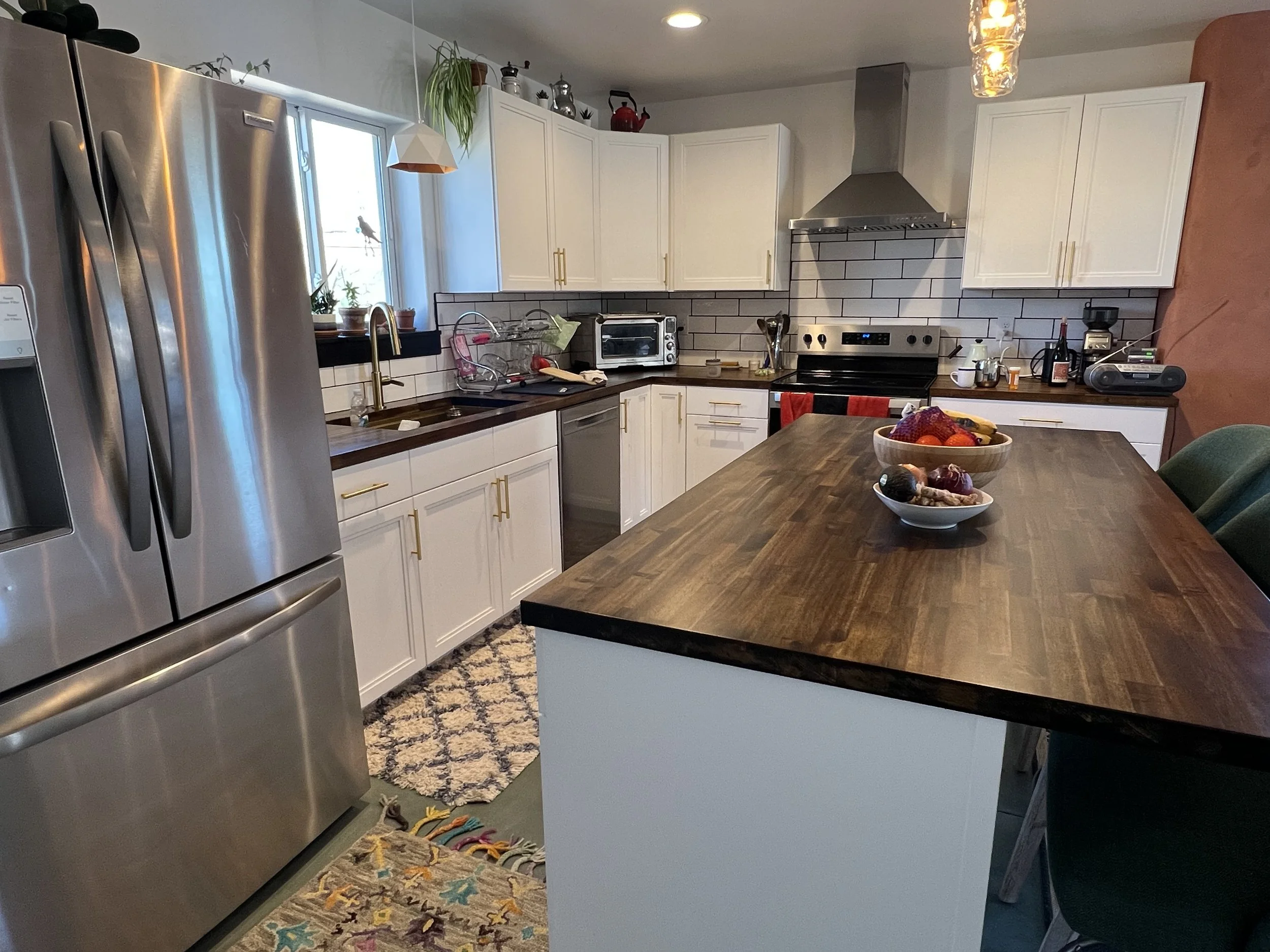 A modern kitchen with white cabinets, a stainless steel refrigerator, a wooden countertop, and a window showing the outdoors. The kitchen has various appliances, a fruit bowl on the table, and decorative plants.