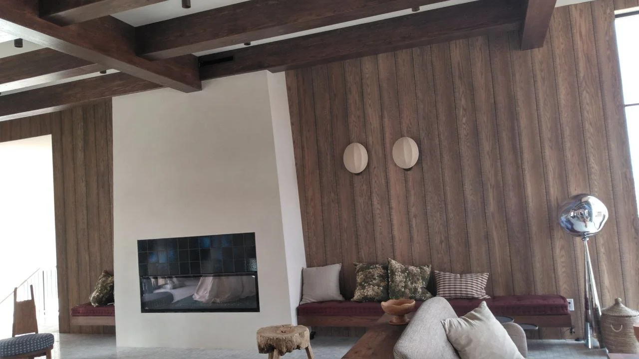 A contemporary living room with a wood-paneled accent wall, a fireplace, and built-in seating with cushions. The room features a mix of modern and rustic decor, including a large metallic floor lamp and decorative items on the coffee table.