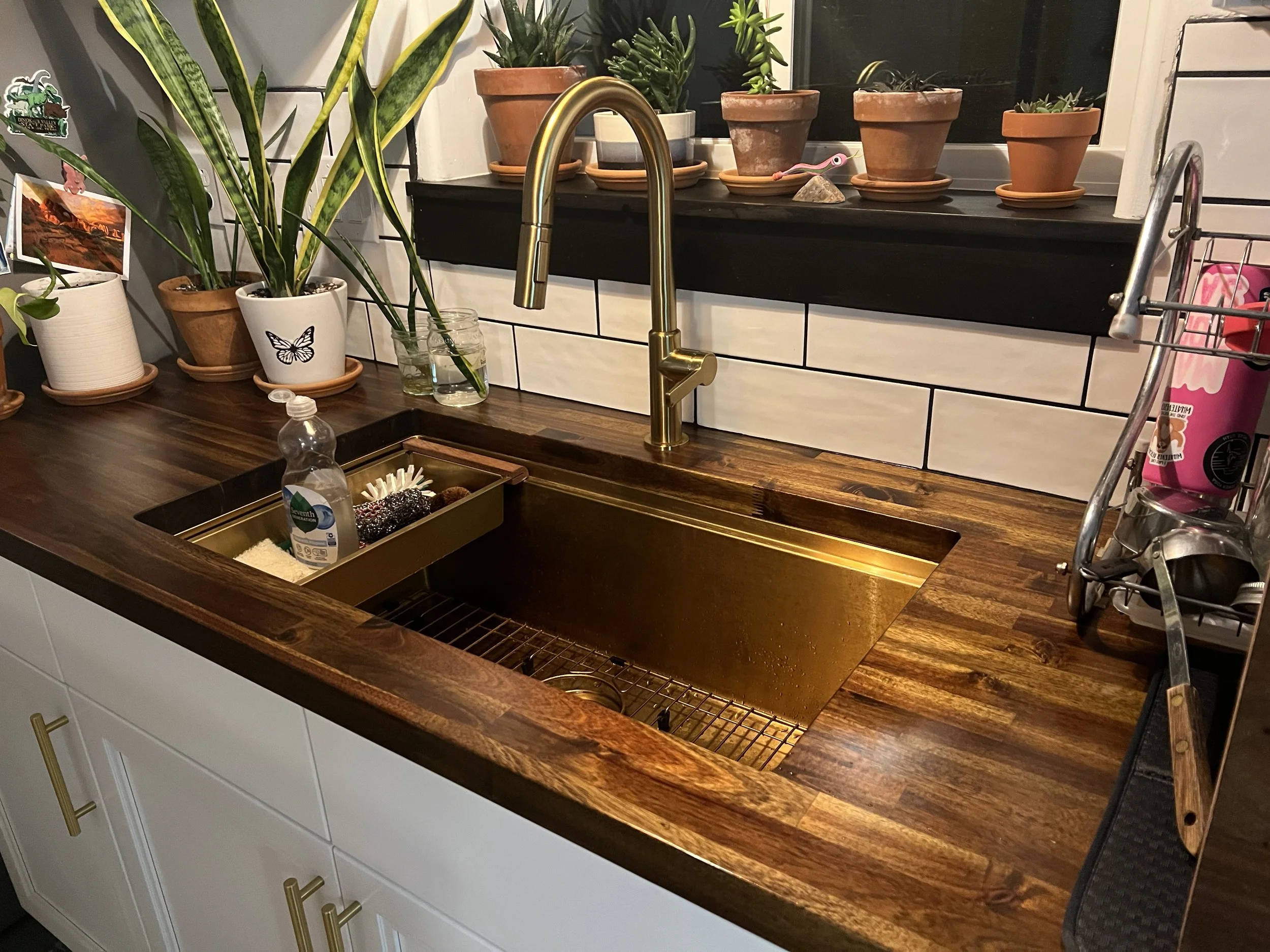 A kitchen countertop with a copper-colored sink, a gold-colored faucet, and white cabinets with gold handles. Several potted plants are placed on a black windowsill and a wooden shelf above the sink. There are also small decorative items and a dish scrubber near the sink.