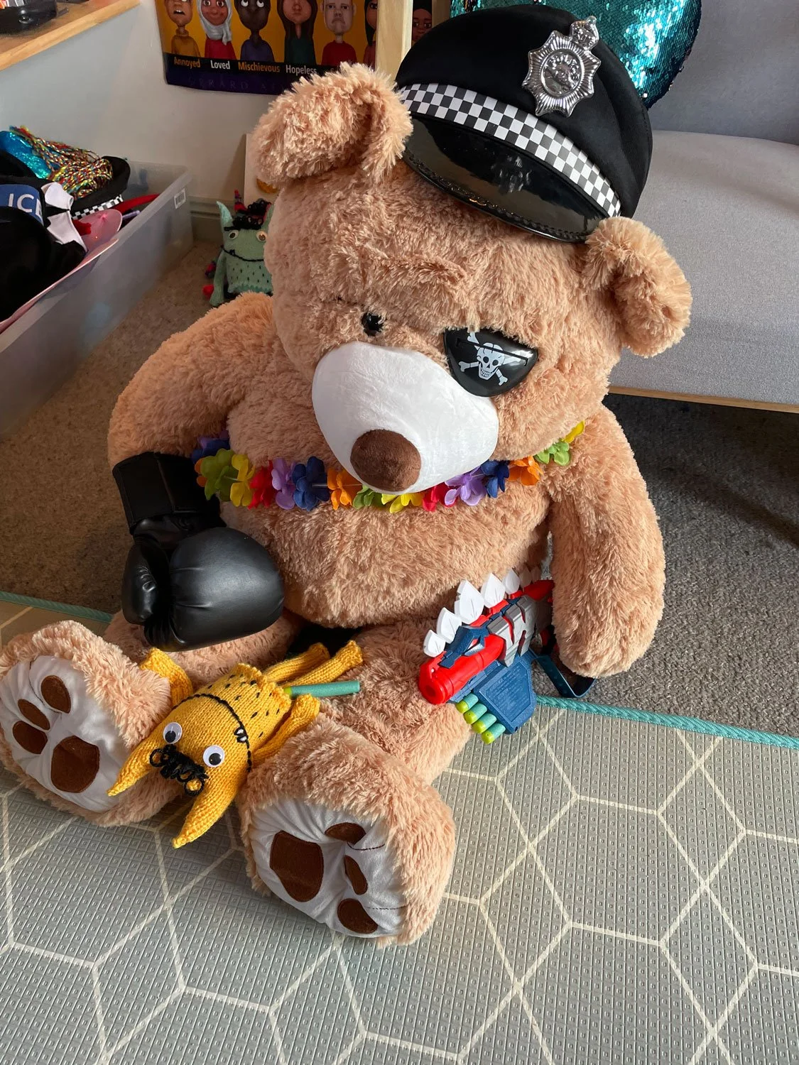 A large teddy bear dressed as a police officer with a police cap, eye patch, and a black glove on one paw. It has a sheriff badge on the cap, a colorful flower necklace, and a toy gun. The teddy bear also has plush banana-shaped slippers on its feet. It is sitting on a textured surface with a background that includes a poster, a drawer with various items, and a small green plush toy.