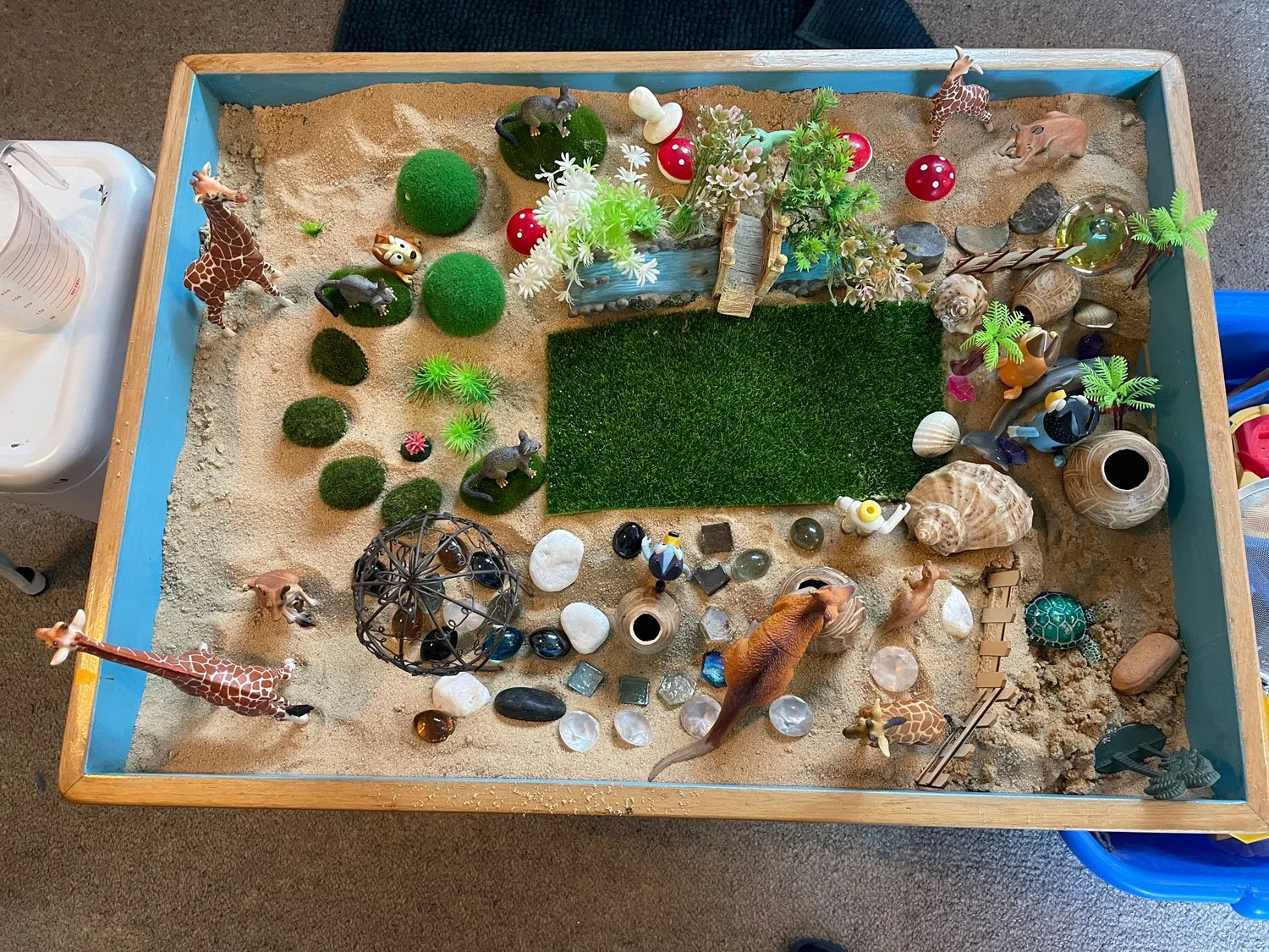 A miniature zoo or safari diorama with toy animals, plants, and decorative items on a sand and grass surface in a wooden tray.
