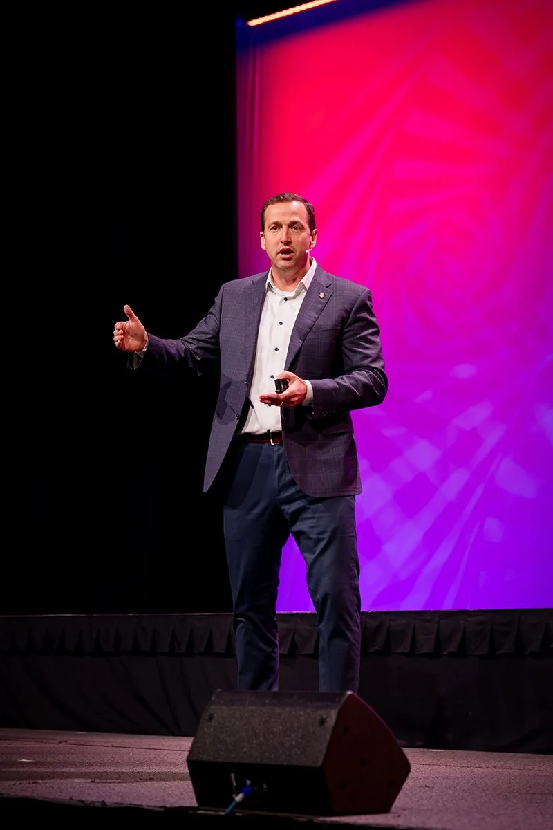 A man in a suit and white shirt giving a presentation on a stage with a colorful pink and purple background.