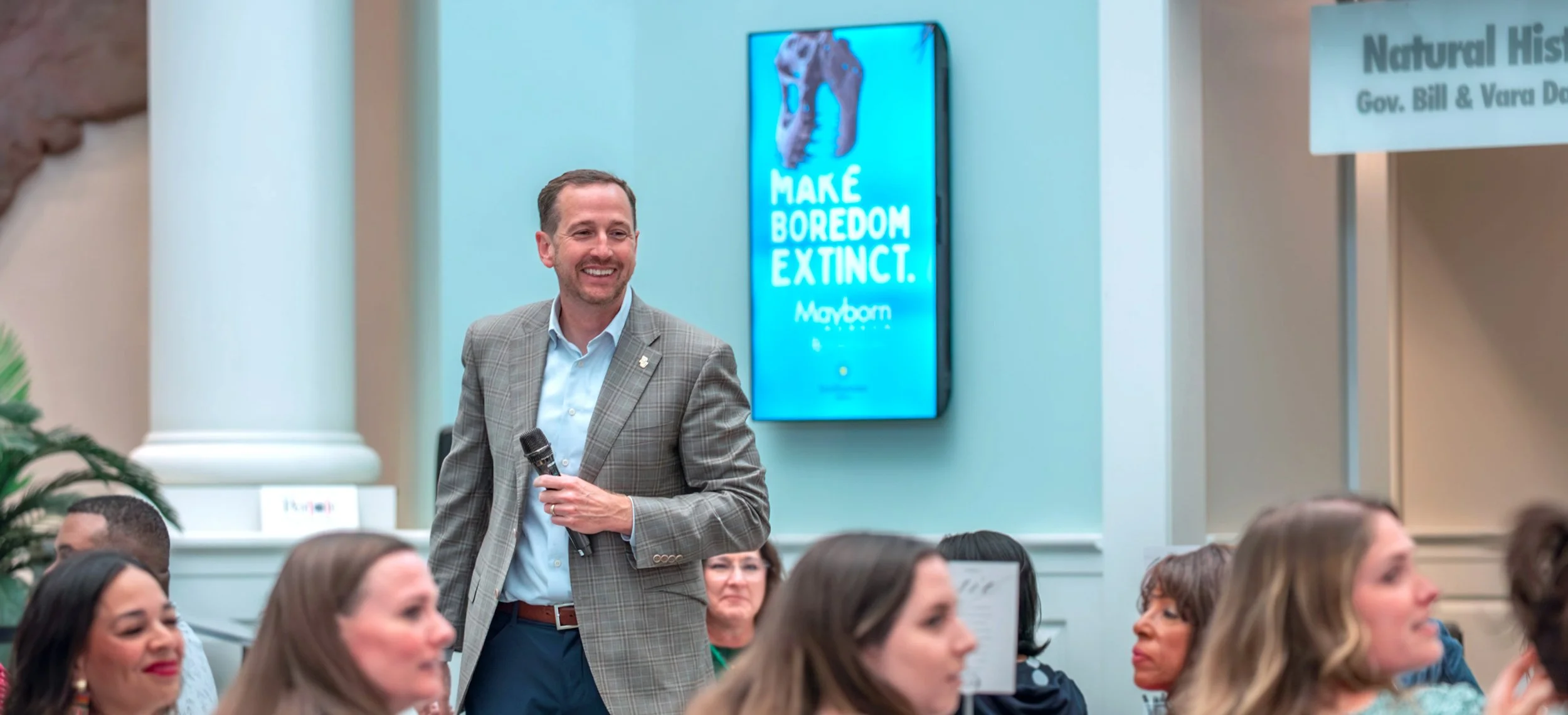 A man in a gray plaid suit holding a microphone smiling at a gathering of people in a room with a blue digital display behind him that says "MAKE BOREDOM EXTINCT."
