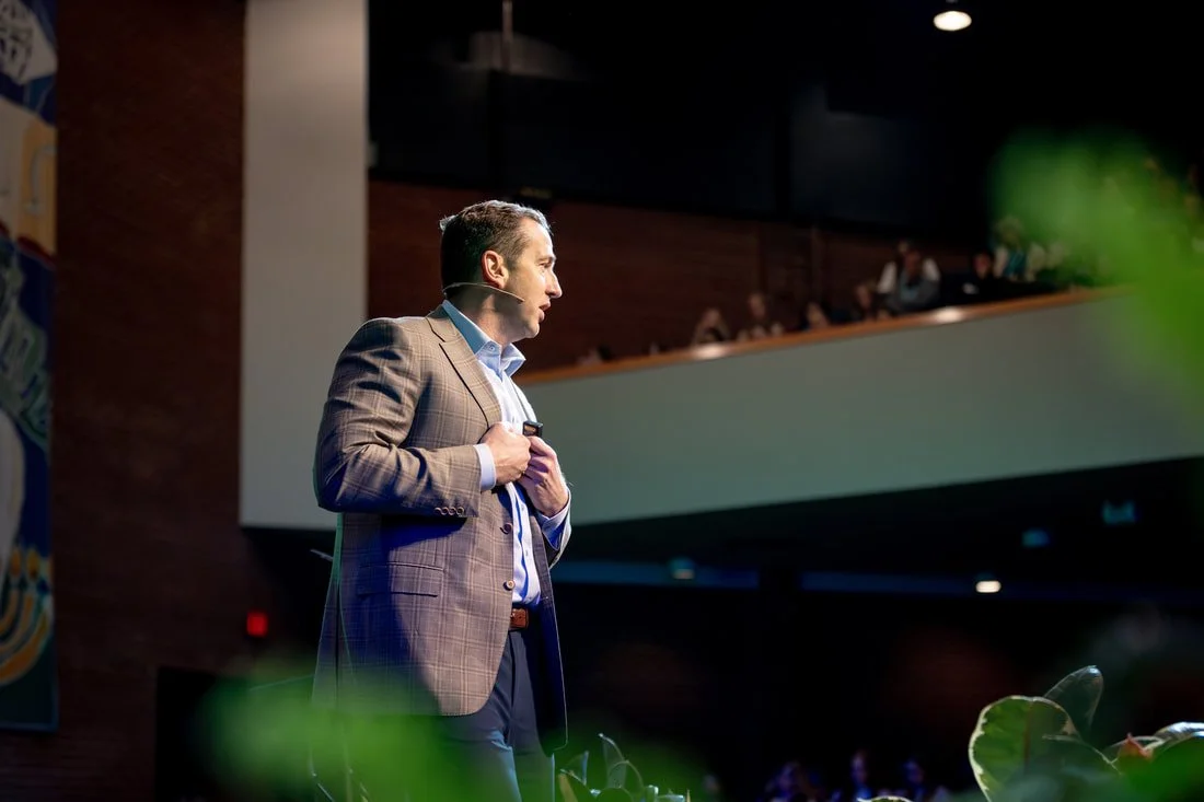 A man in a brown plaid blazer giving a presentation on stage with a microphone headset